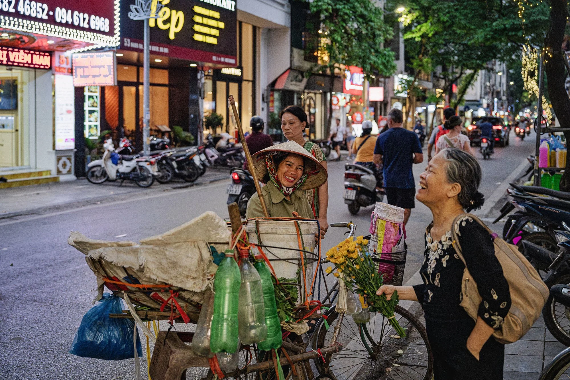 one woman with gardening tools on a bicycle and one woman holding a bouquet laughing together on the street in hanoi vietnam