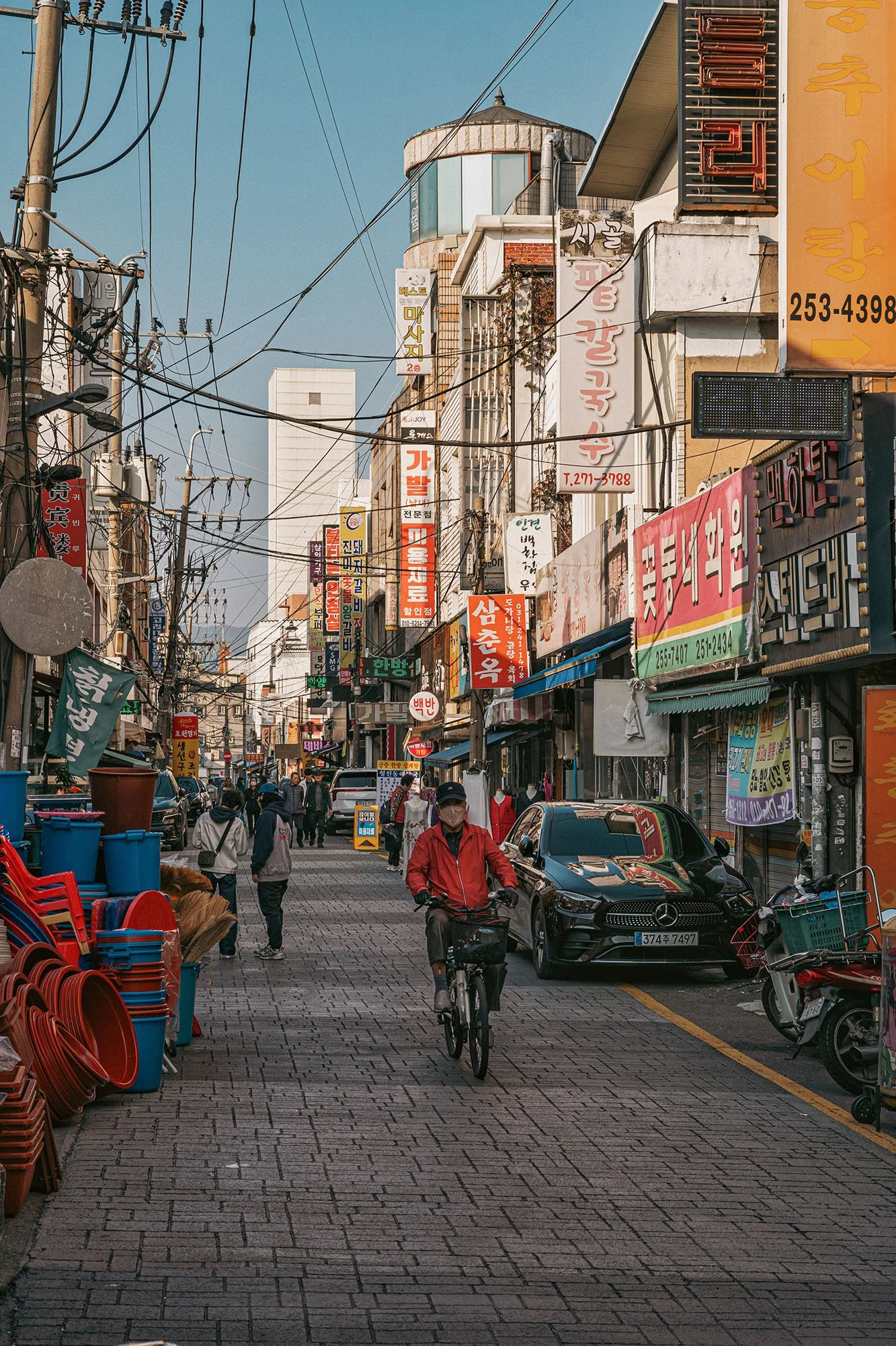 man wearing mask and red jacket riding a bicycle on an old shopping street with wires overhead in suwon korea