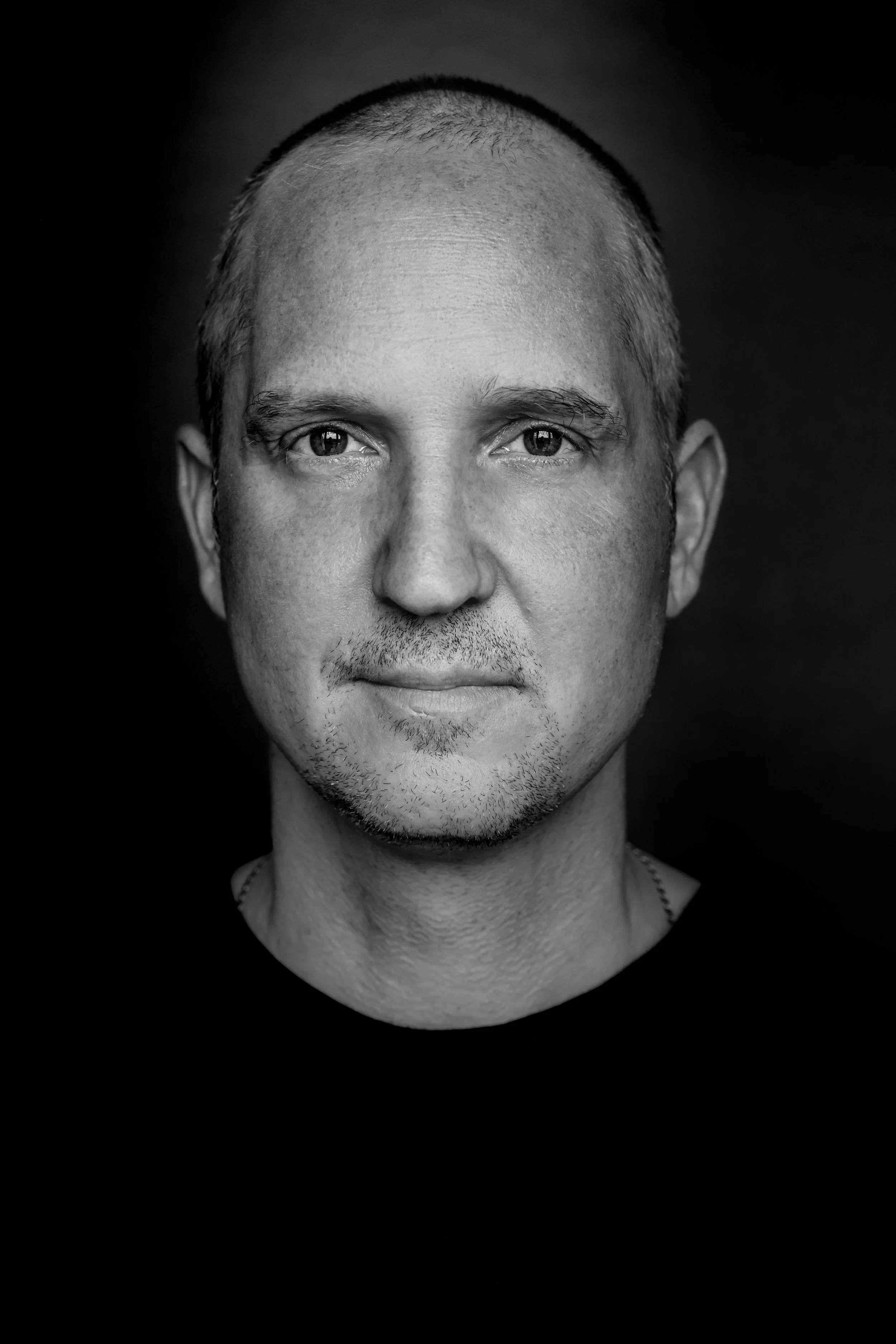 Kontrastrecihes schwarz-weiß Portrait von Volker Walzer mit kurz geschnittenem Haar, eher heller Haut, kurzem Bart und einem leichten Lächeln vor dunklem Hintergrund.