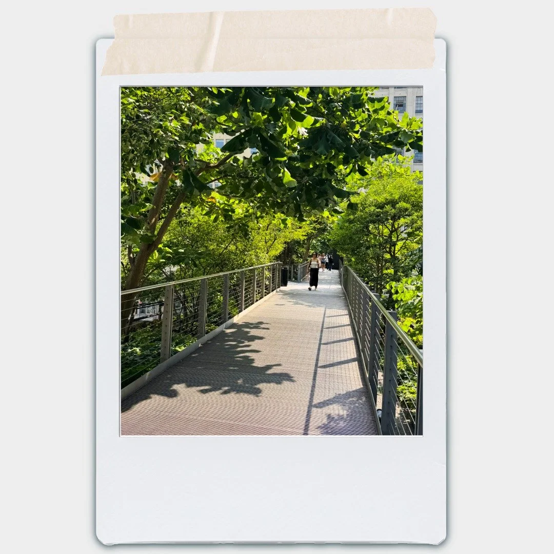 A walkway surrounded by green trees and foliage, with a woman walking away in the distance, and a cityscape visible in the background.