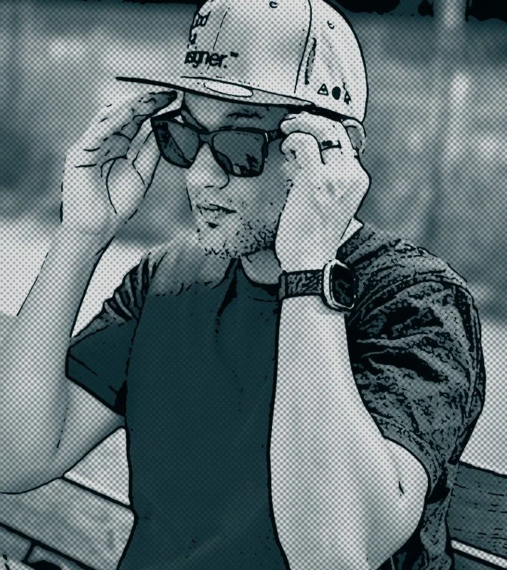 A man wearing a baseball cap and sunglasses adjusting his glasses outdoors.