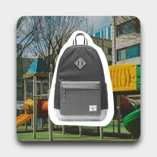 Small black backpack with front pocket, placed in front of a playground with slides and climbing equipment, trees, and buildings in the background.