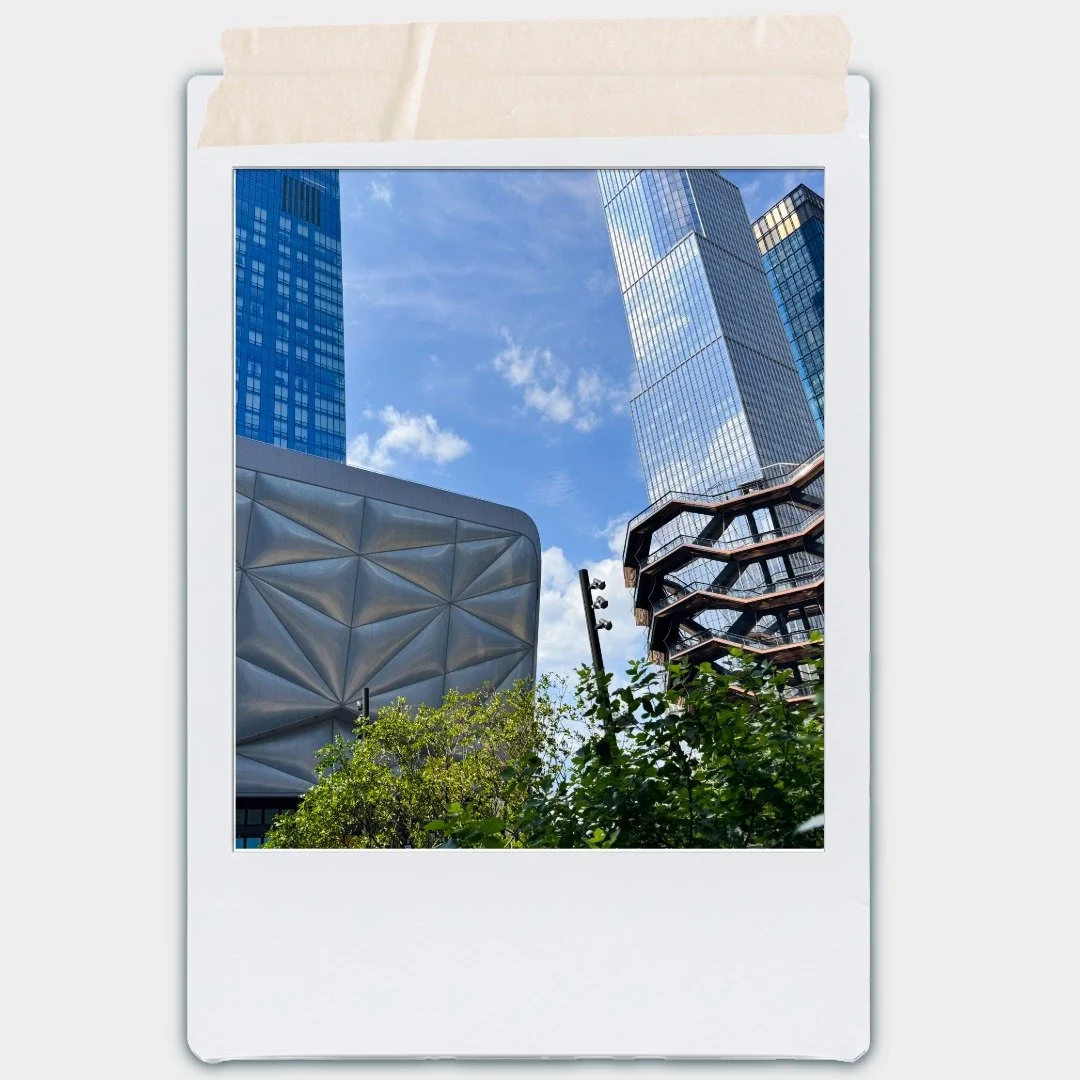 Cityscape with modern high-rise buildings, a unique metallic building, and a glass tower under a blue sky with clouds, framed by greenery in the foreground.
