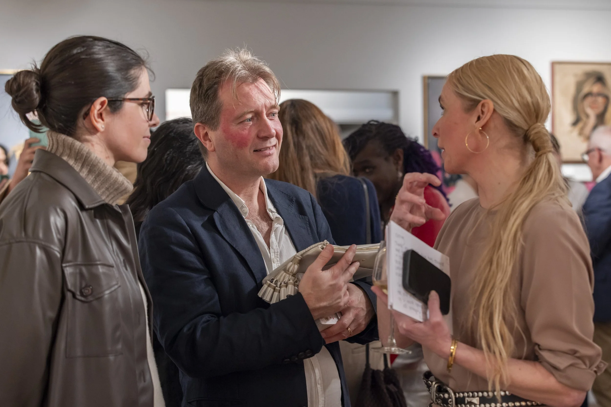Three people engaged in a conversation at a social gathering, with artwork hanging on the wall in the background.