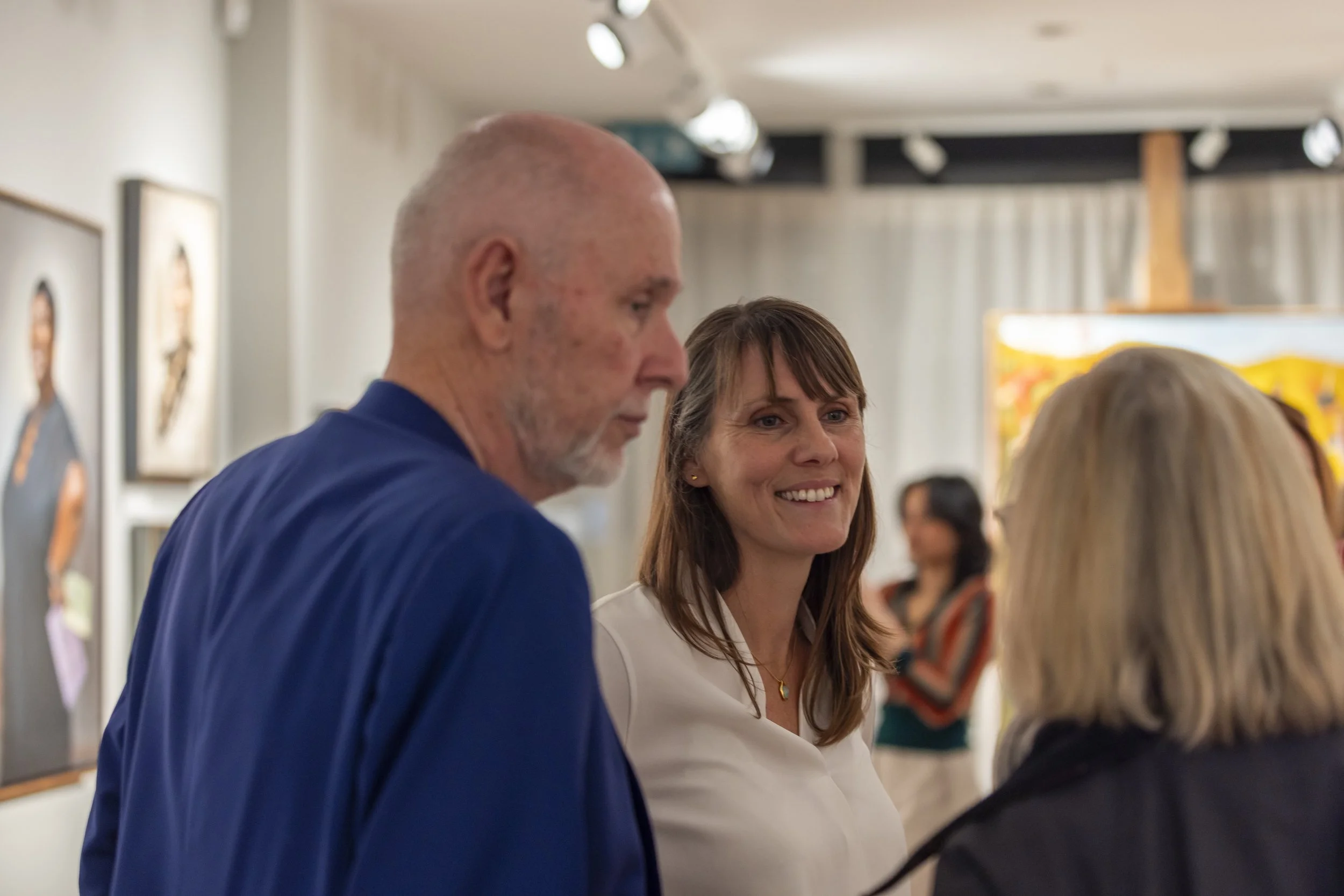 Three people talking in an art gallery, with paintings visible on the walls behind them.