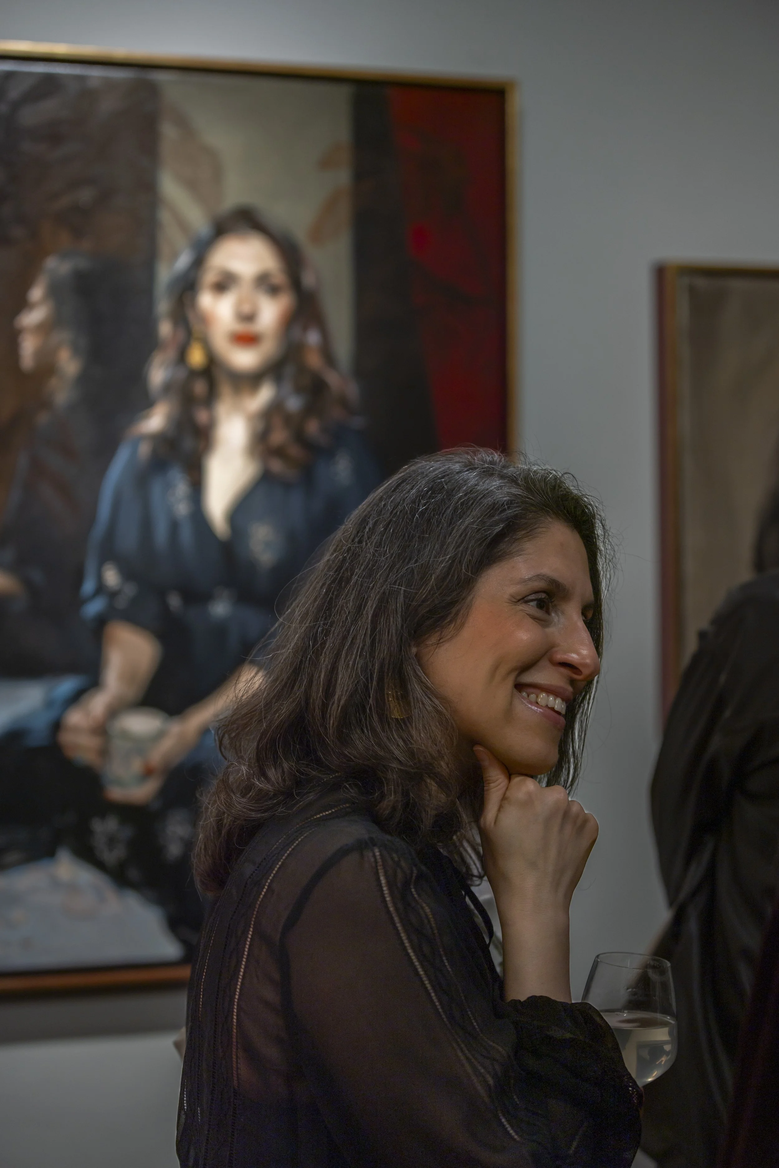 A woman smiling while holding a glass of white wine at an art gallery, with a portrait painting in the background.