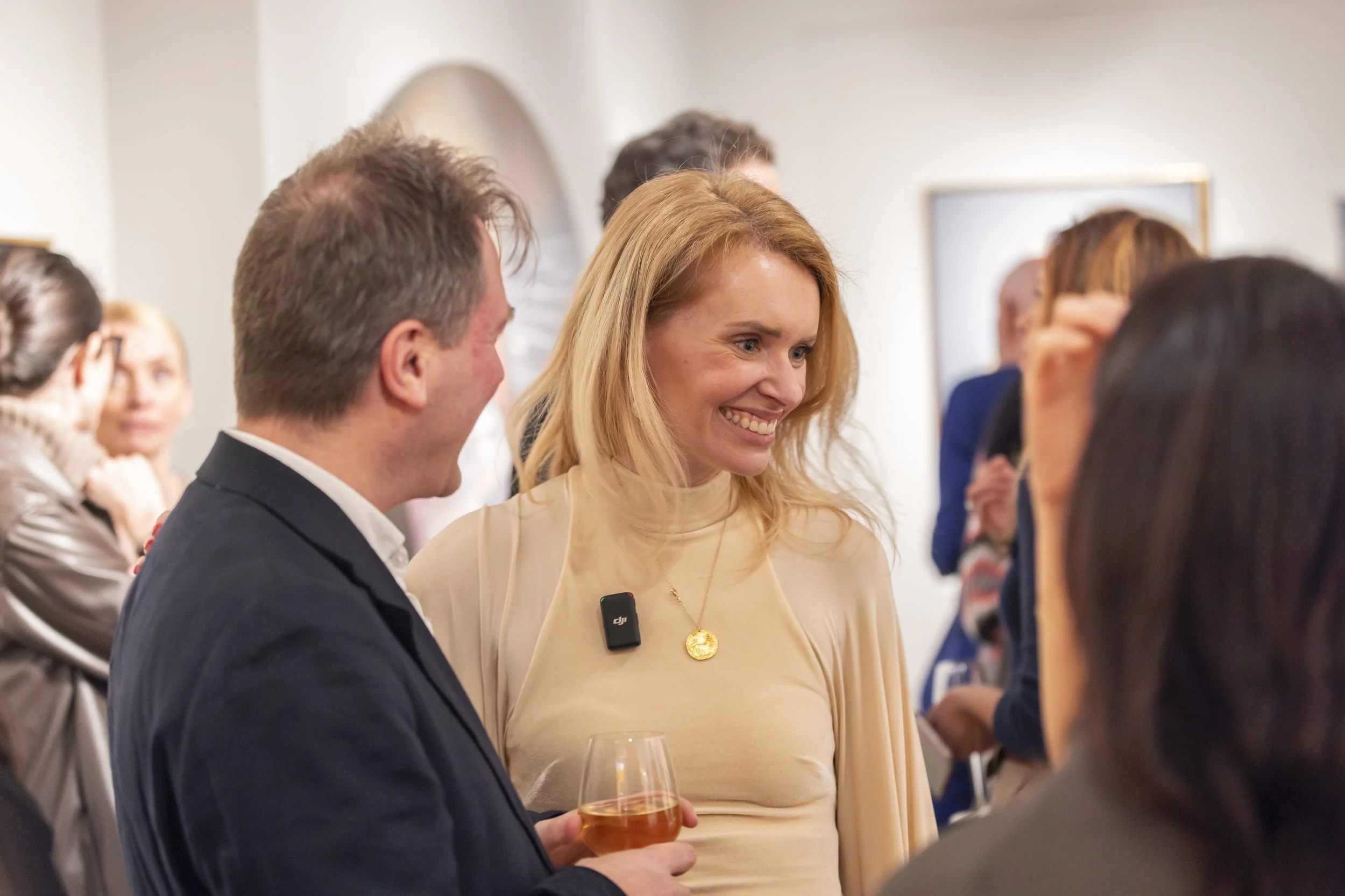 People socializing at an indoor event, including a woman with blonde hair in a beige top and a man in a suit holding a glass of wine.