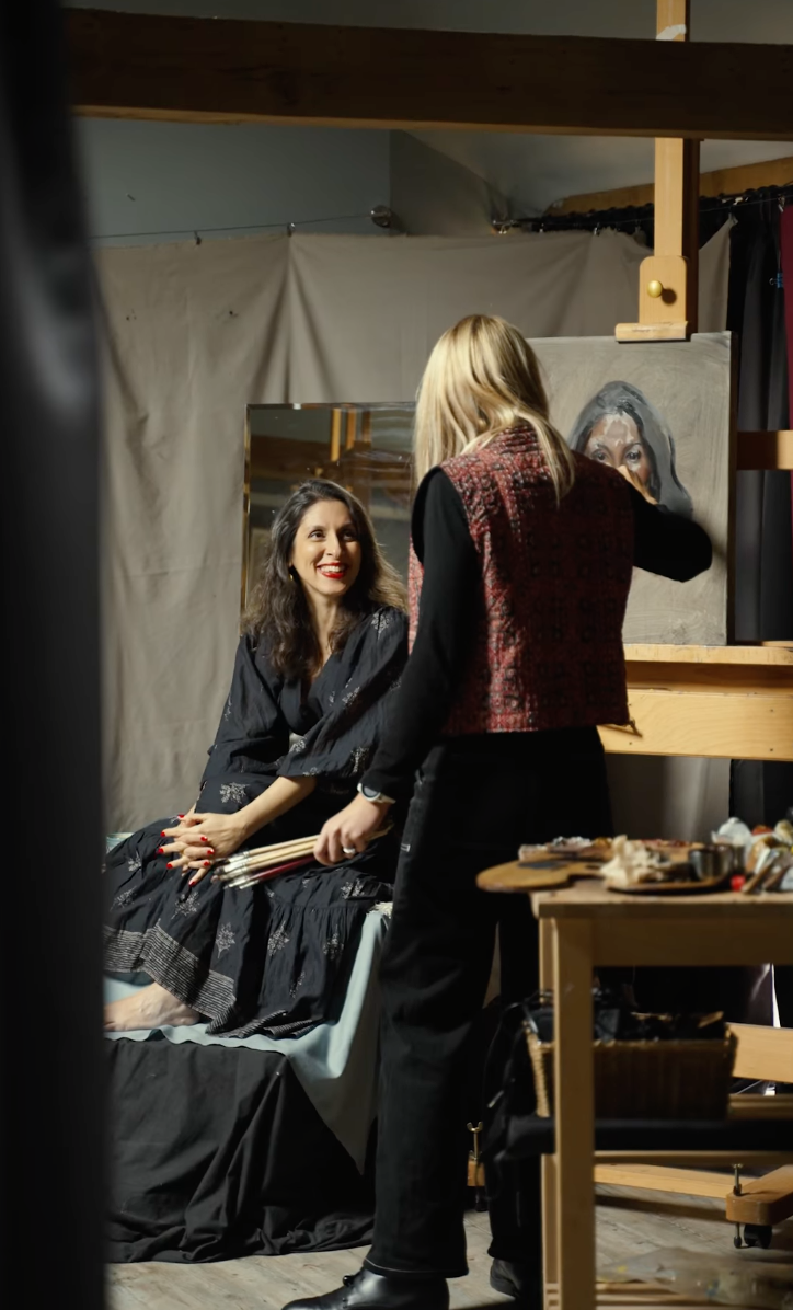 A woman is painting a portrait of another woman on an easel in an art gallery or studio. The painter is smiling at the camera, and the woman being painted is seated nearby. The background features various paintings and bookshelves.
