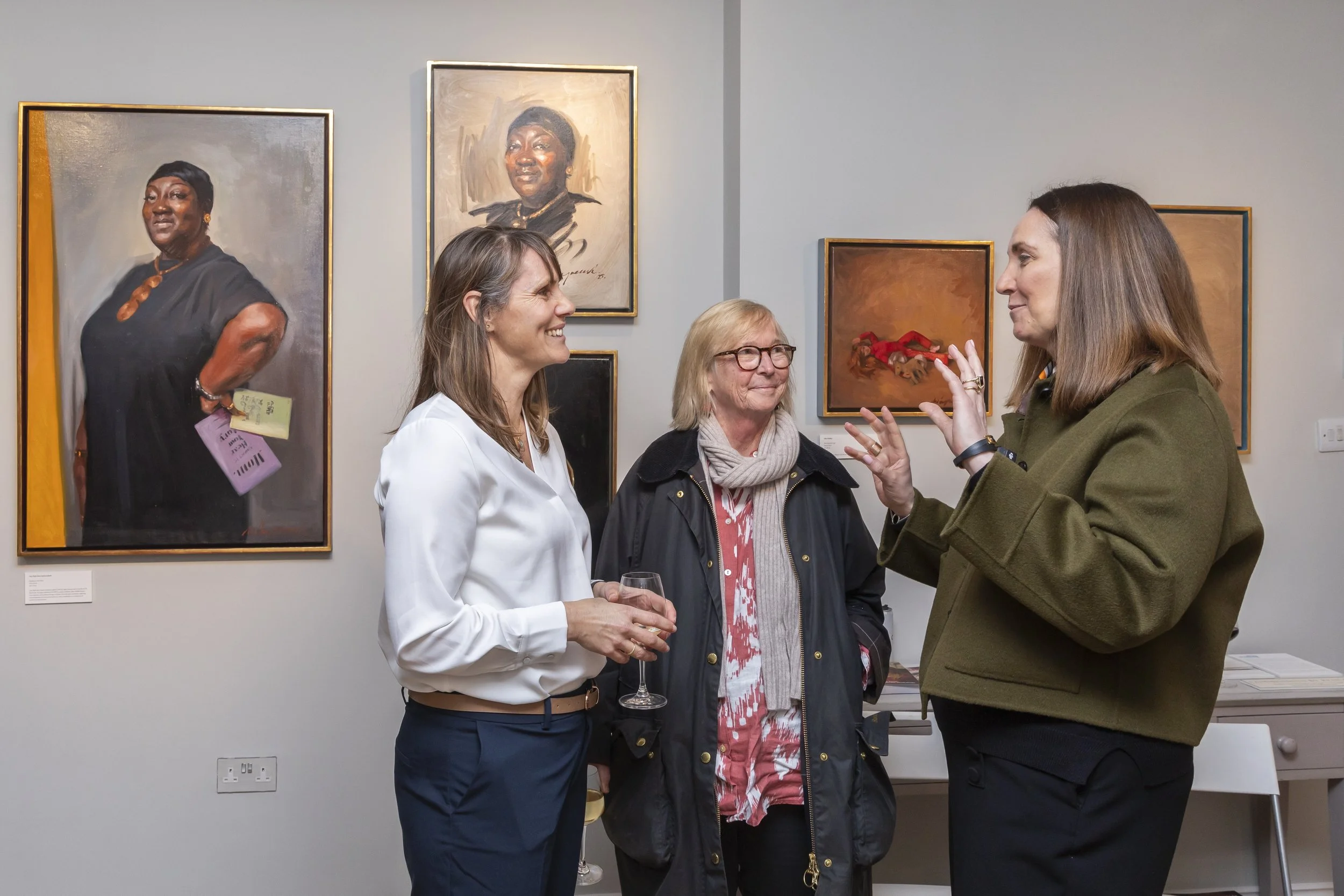 Three women in conversation at an art gallery, with paintings of women on the wall behind them.