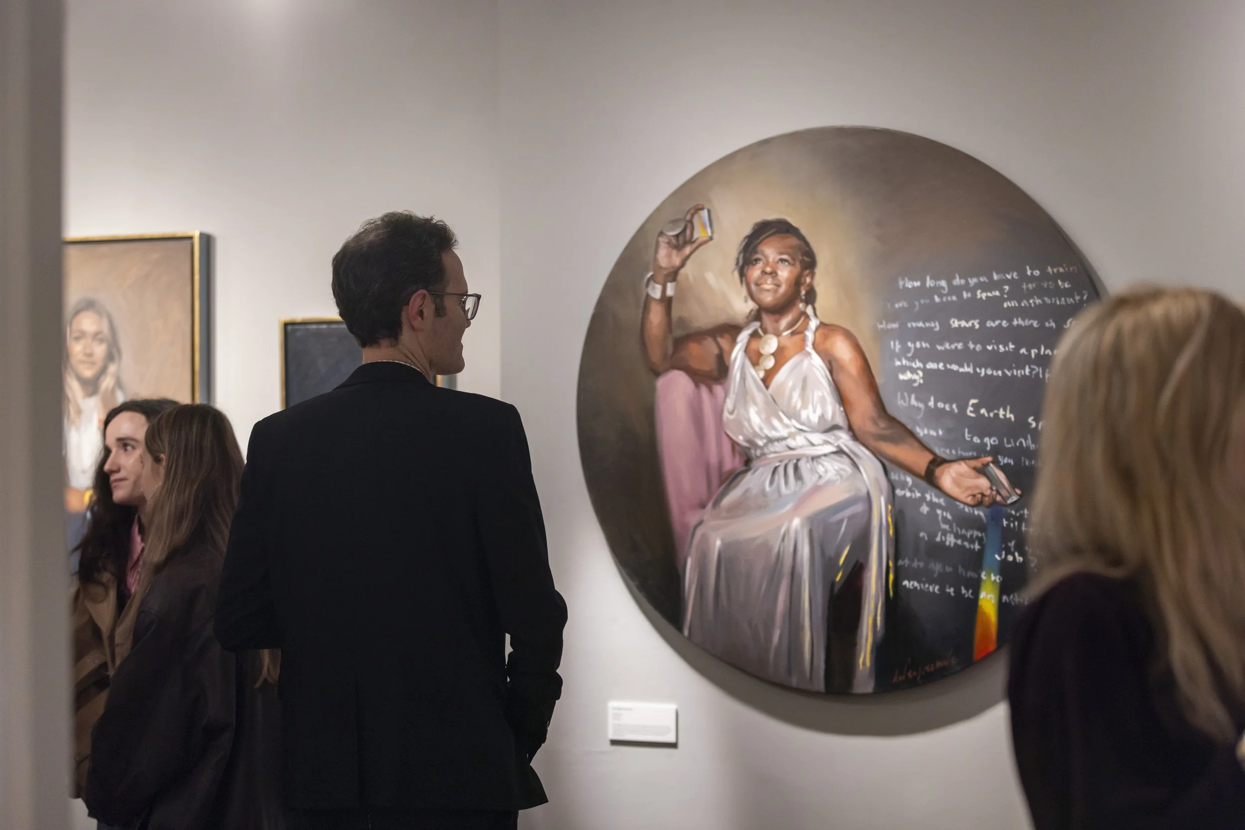 People in an art gallery viewing a circular painting of a woman taking a selfie, with other framed artworks on the wall.