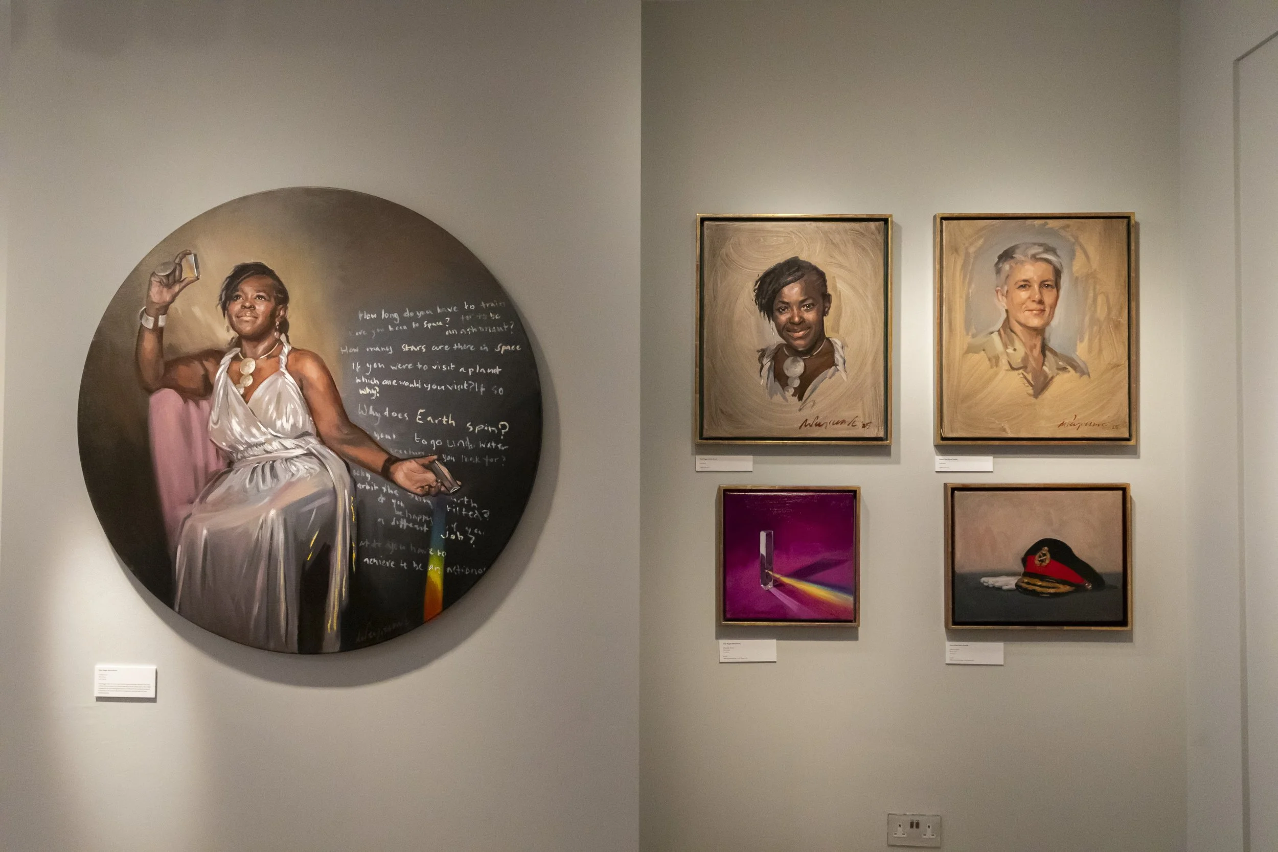Art exhibit featuring a large circular portrait of a woman in a white dress holding a phone, with handwritten text around her, and four smaller portraits on the adjacent wall including two women, a stylized abstract piece with a purple background, an