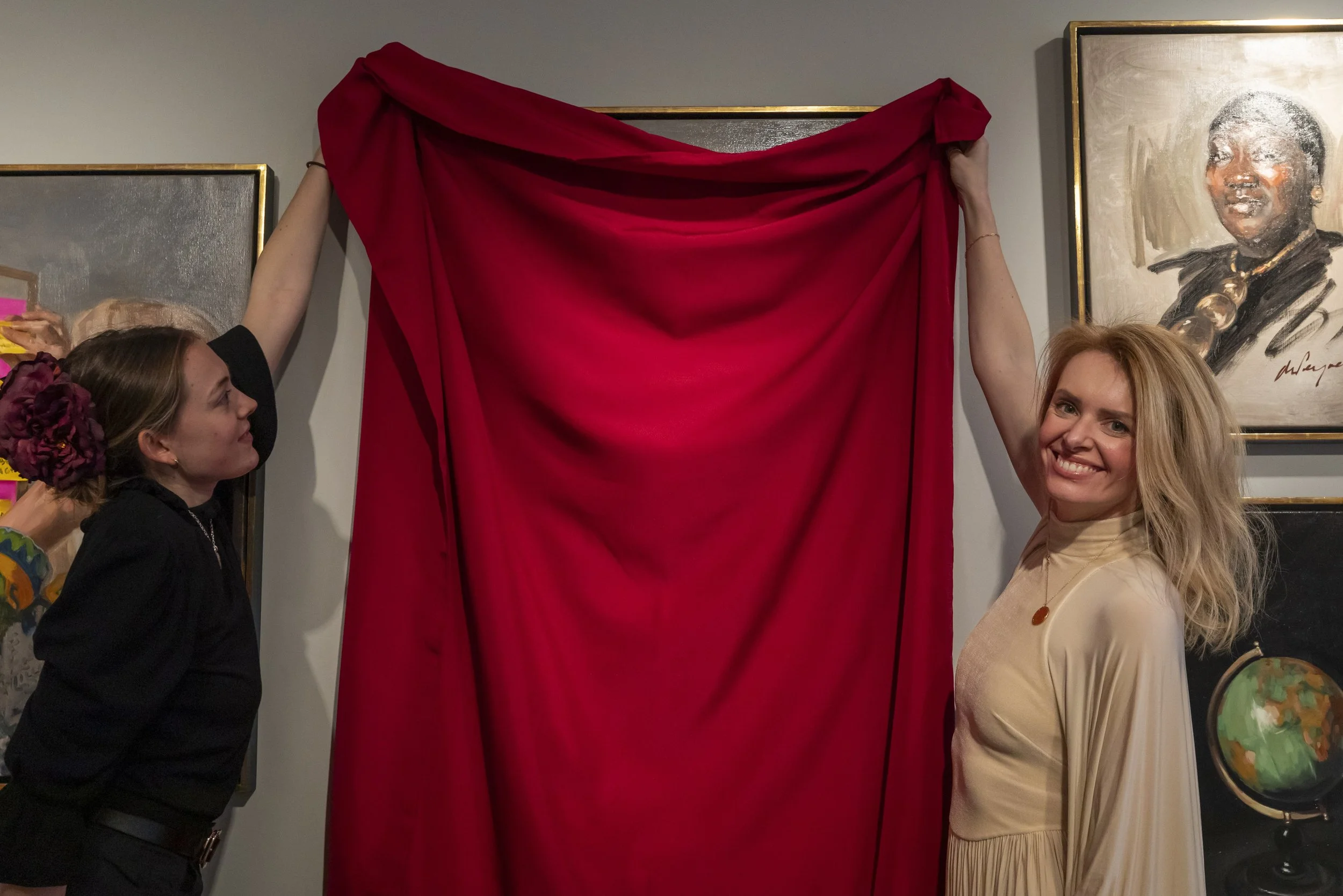 Two women unveiling a red curtain in an art gallery, with paintings on the walls behind them.