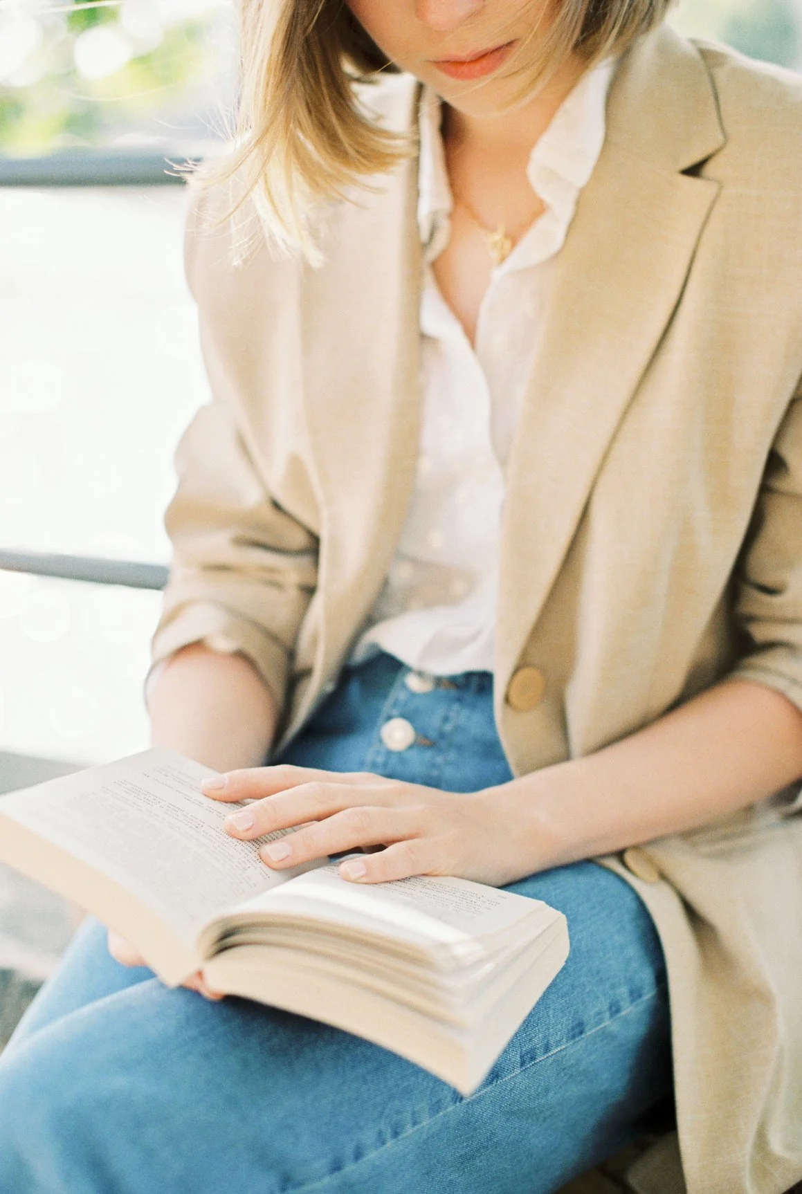 image of a blonde woman in a beige blazer and blue jeans reading a book outside
