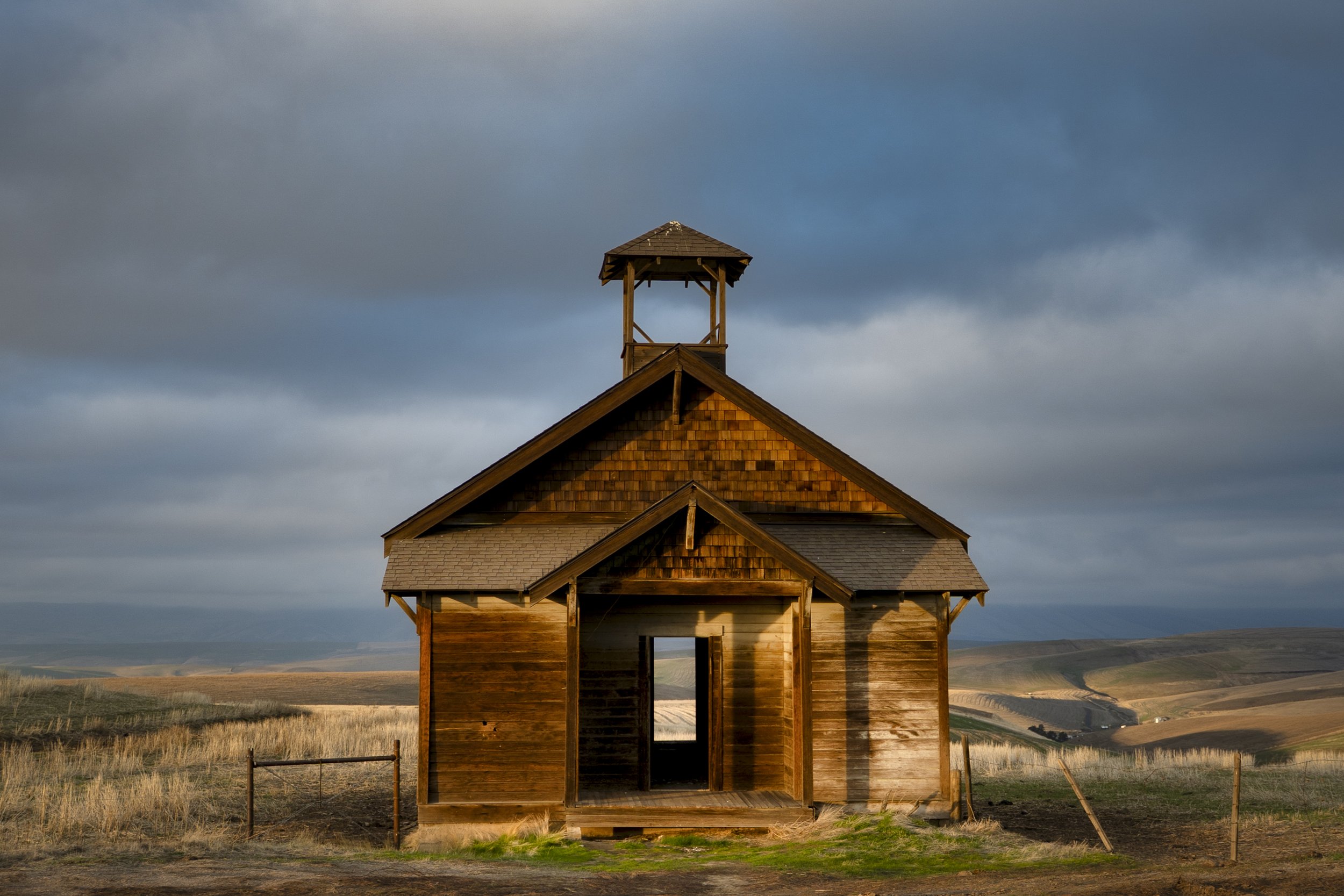 A wooden church with a bell tower on a grassy landscape under a cloudy sky.