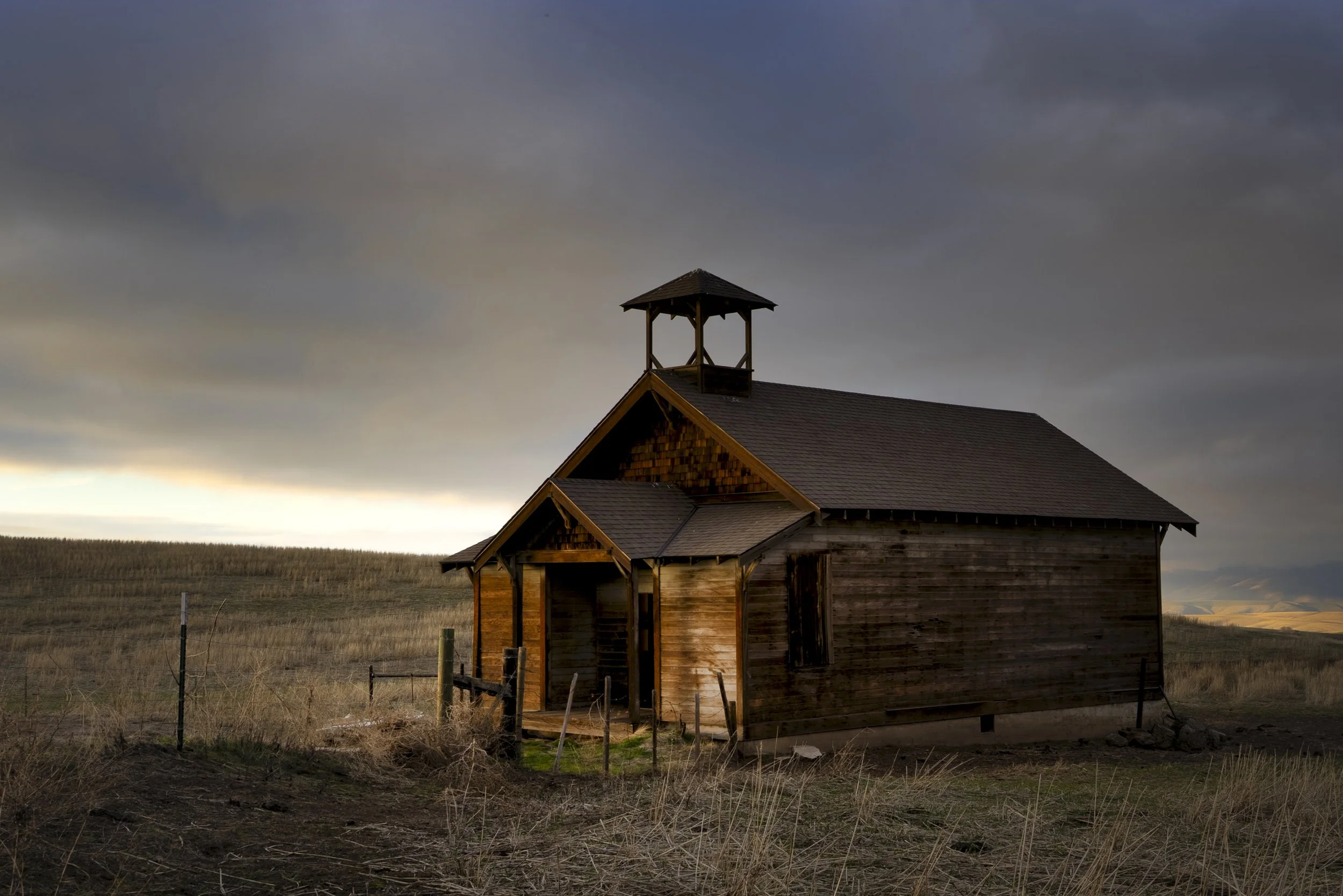A small, rustic wooden church in a rural field at sunset, with dark clouds overhead.