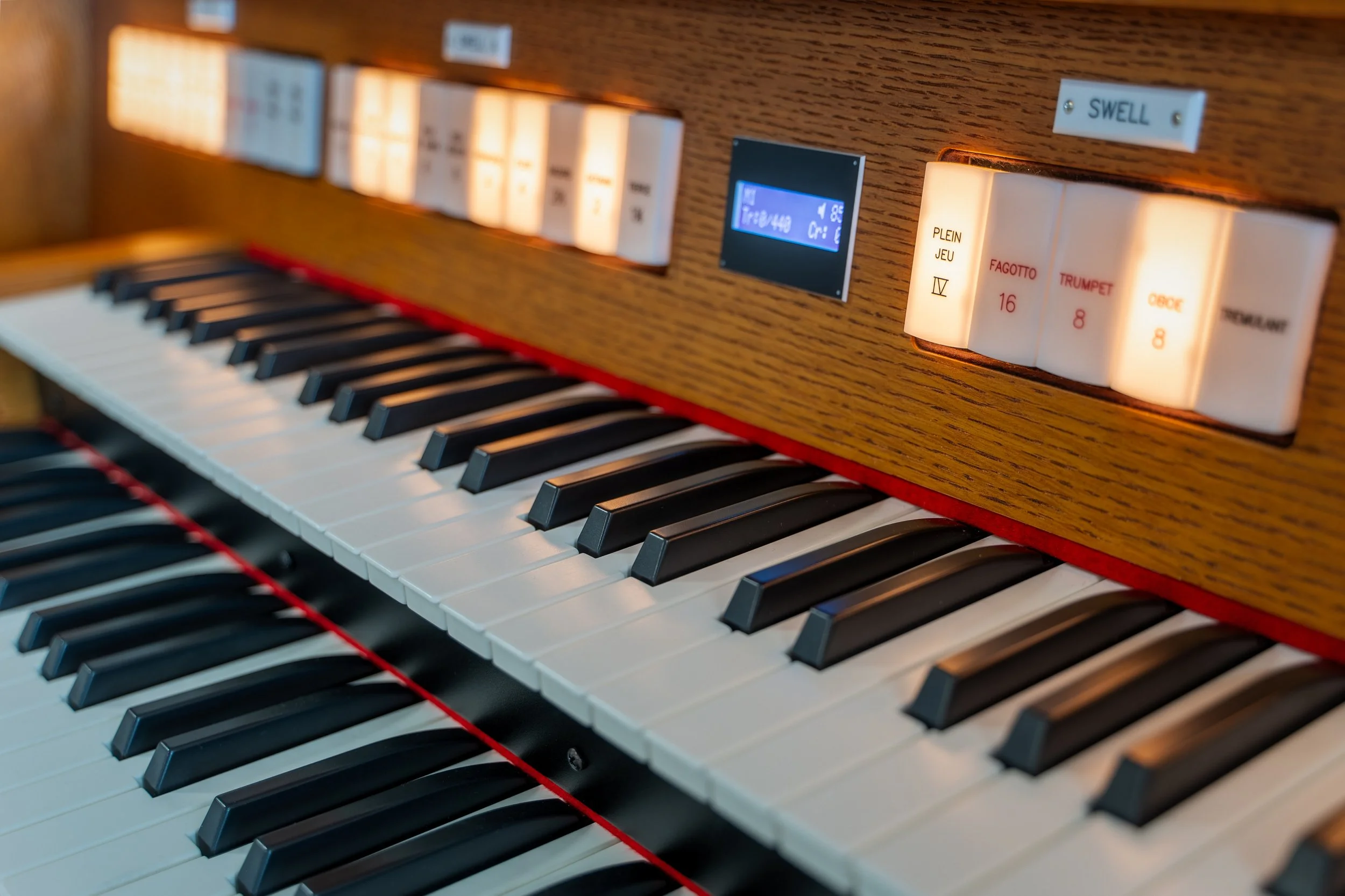 Close-up of an electronic organ keyboard with white and black keys, a wooden panel, and illuminated stop tabs labeled with musical terms and numbers.