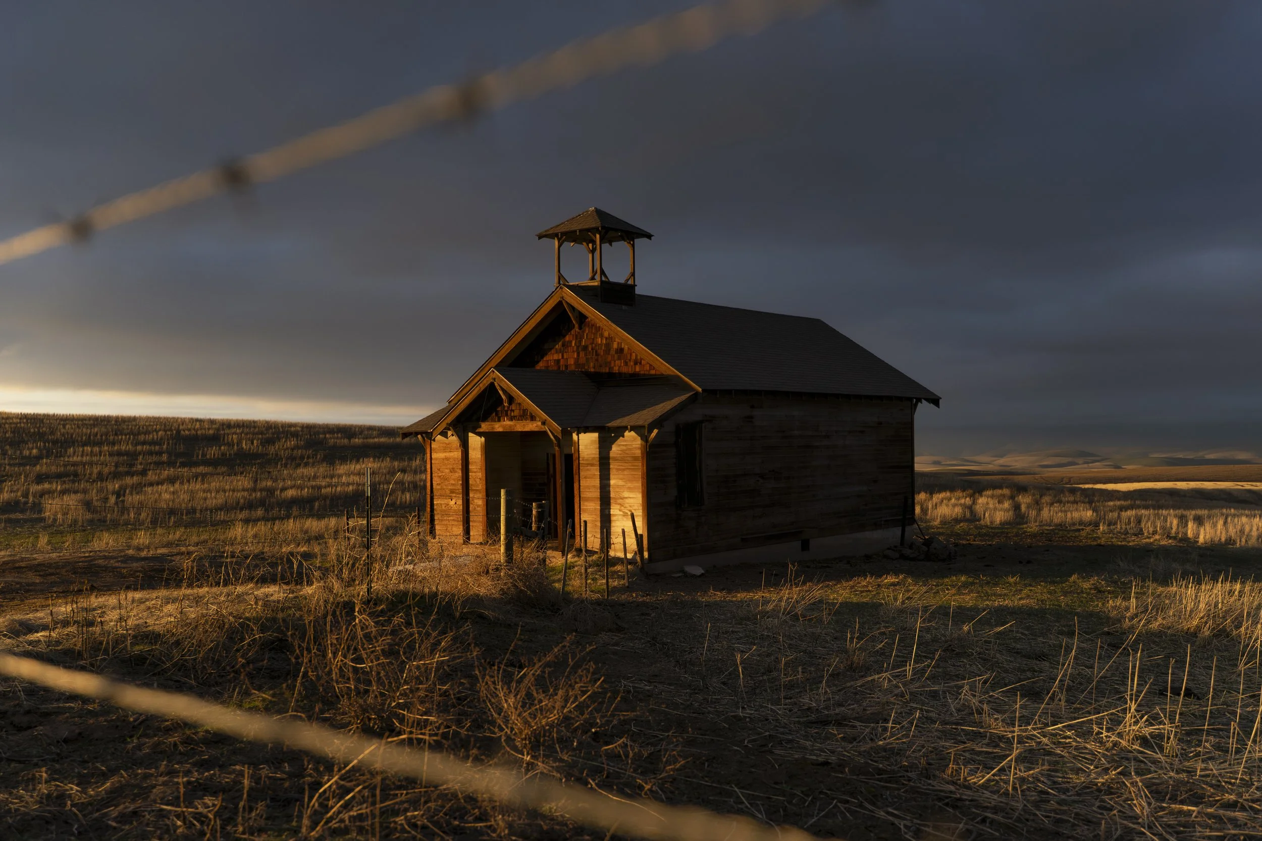 An old wooden church in a rural field during sunset, with dark stormy clouds overhead and a barbed wire fence in the foreground.