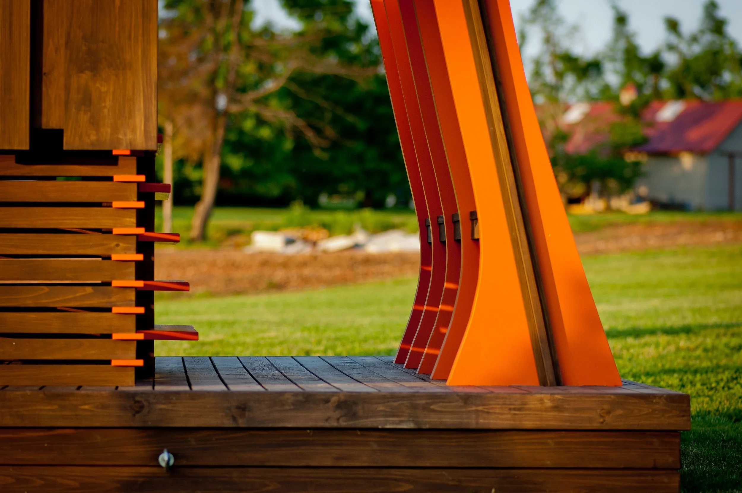 Close-up of a wooden structure with orange metal hanging curved beams, set outdoors on a wooden platform with a grassy field and trees in the background.