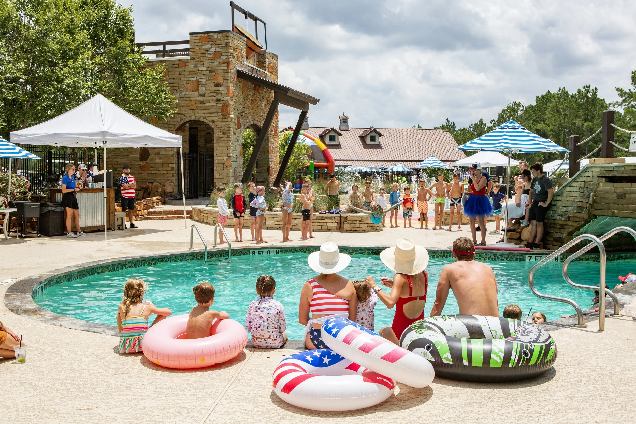 People at a poolside during a summer event, with children and adults, some in swimsuits, sitting or floating on pool inflatables, and a group of children standing in a line near the pool, with some on a stage area with blue and white umbrellas overhe