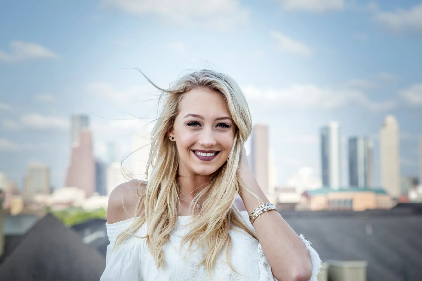 A smiling blonde woman with long hair, wearing a white off-the-shoulder top, standing outdoors against a city skyline with modern skyscrapers and a cloudy sky. Photographed by Houston Photographer Katie Park.