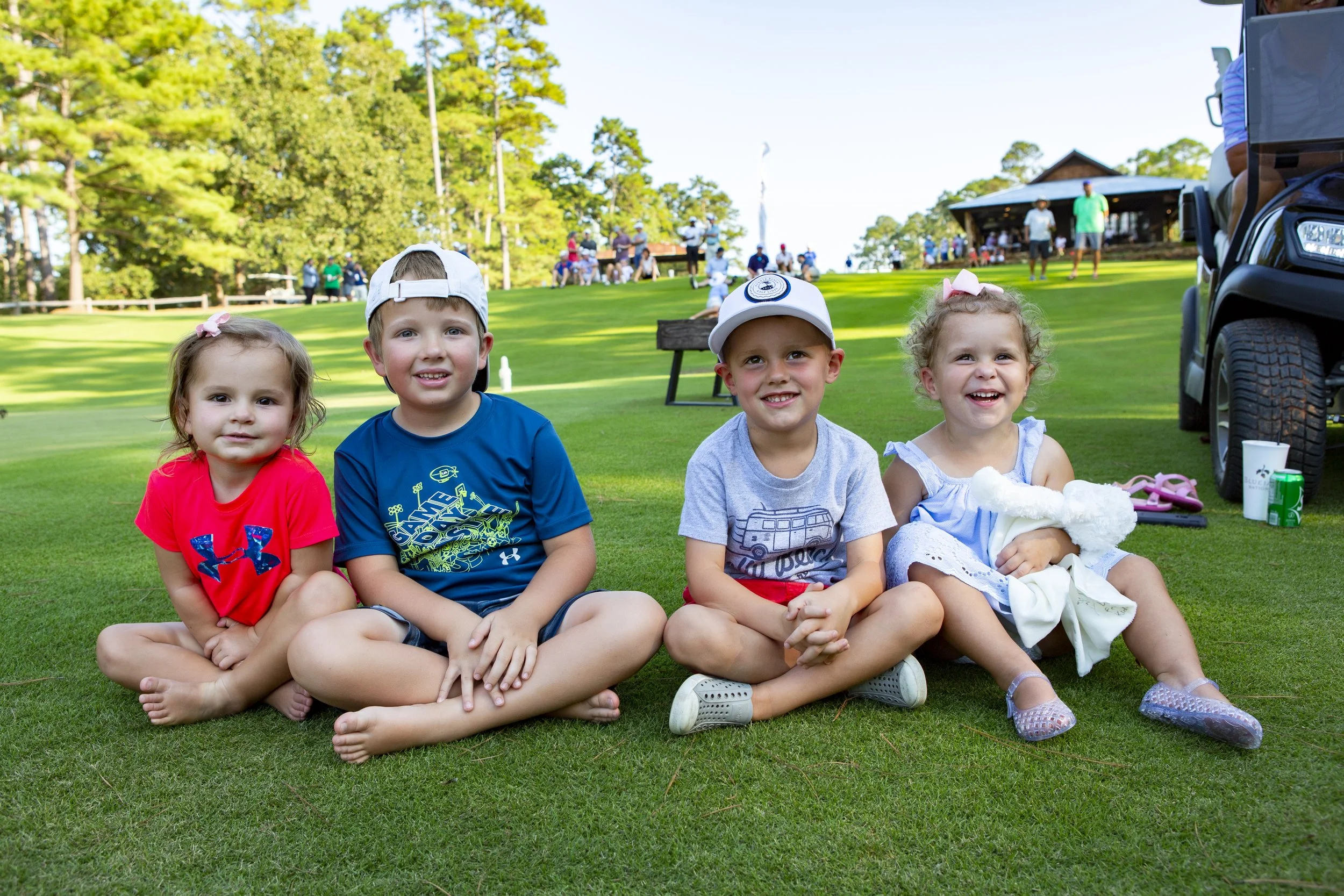 Four young children sitting on grass at a golf course, with trees and people in the background, enjoying sunny day.