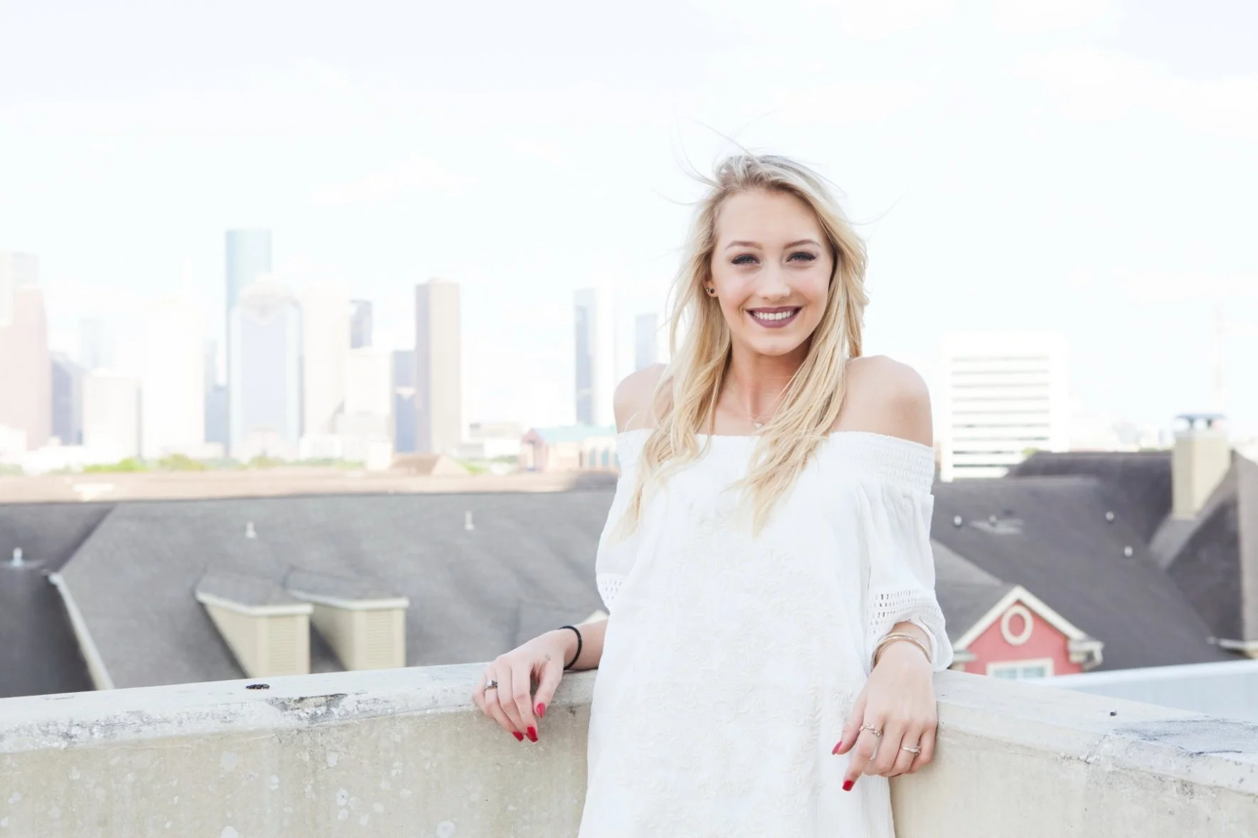 Smiling young woman with long blonde hair wearing an off-the-shoulder white dress, standing outdoors on a rooftop with a city skyline in the background. Photographed by Houston Photographer Katie Park.