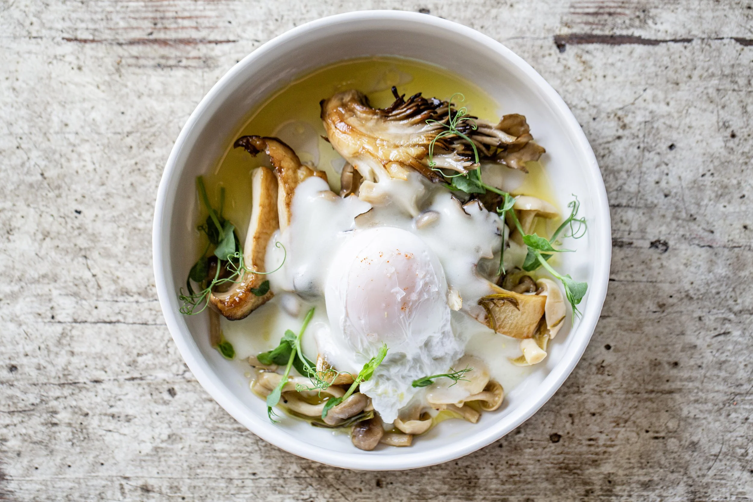 A white bowl containing a soft-boiled egg with a runny yolk, surrounded by sautéed mushrooms, microgreens, and a drizzle of oil, on a rustic wooden surface.