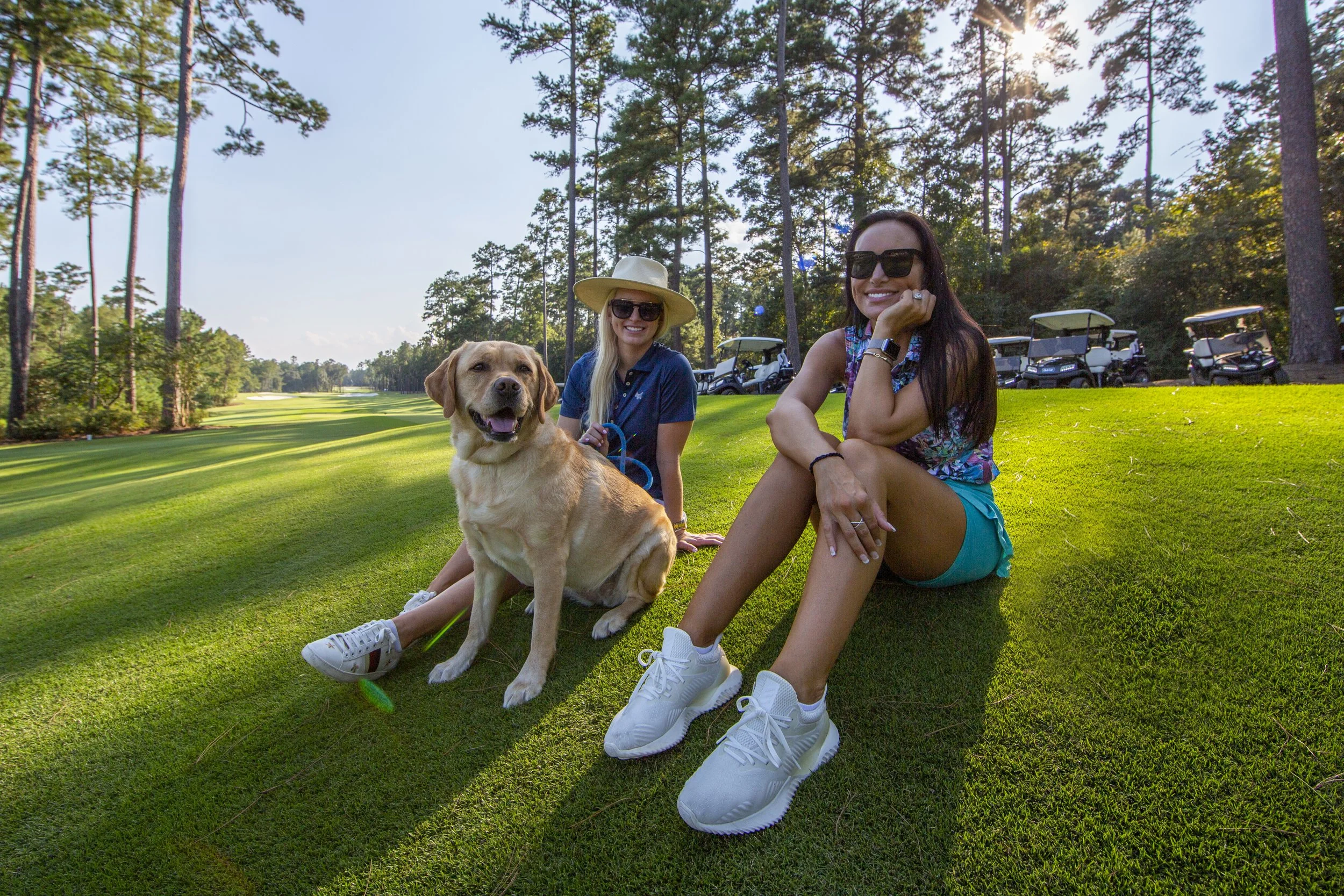 Two women sitting on a golf course with a yellow Labrador Retriever, surrounded by tall trees and golf carts, on a sunny day.