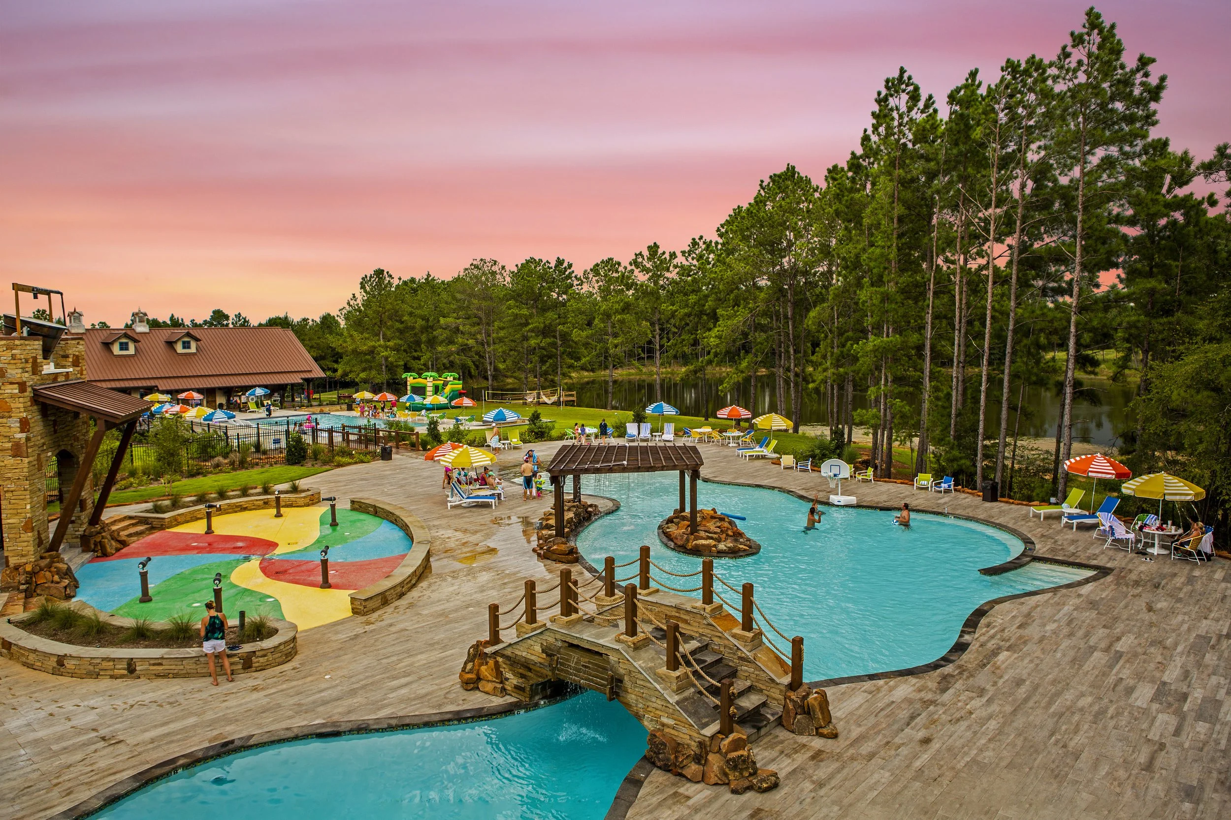 Resort swimming pool area at sunset with colorful umbrellas, lounge chairs, children playing, and a body of water surrounded by trees.