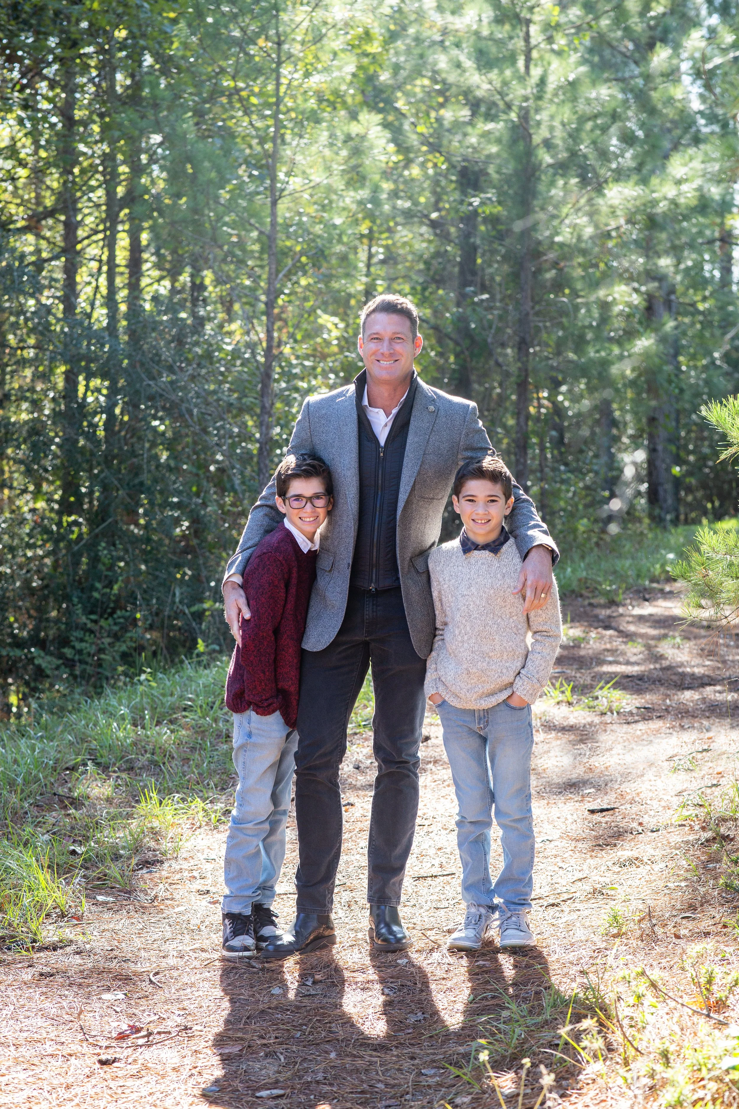 A man and two boys stand on a wooded trail, smiling, with sunlight filtering through trees. Photographed by Houston Photographer Katie Park.