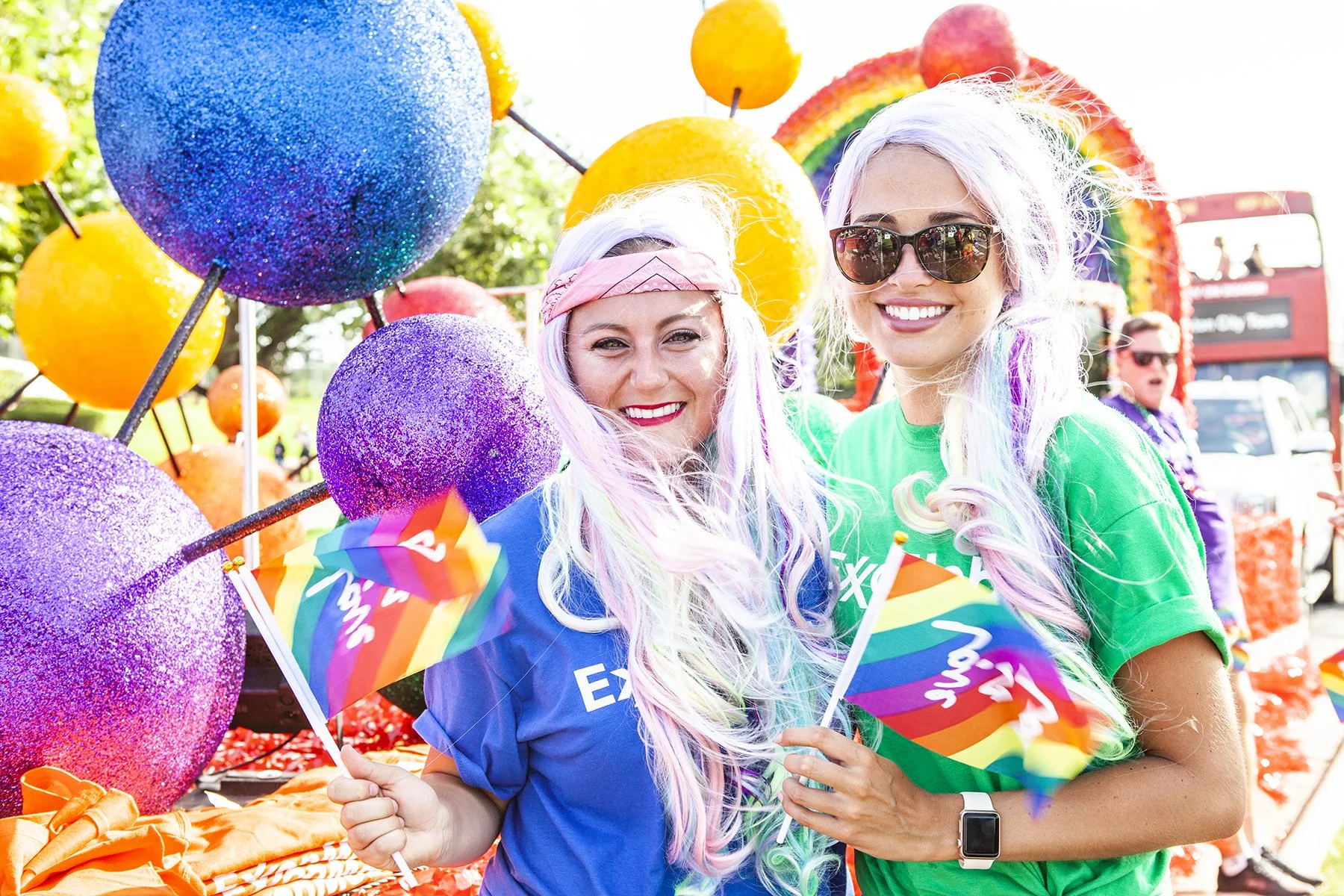 Two smiling women with colorful wigs and sunglasses holding rainbow pride flags at an outdoor pride event, surrounded by large glittery balloons and rainbow decorations.