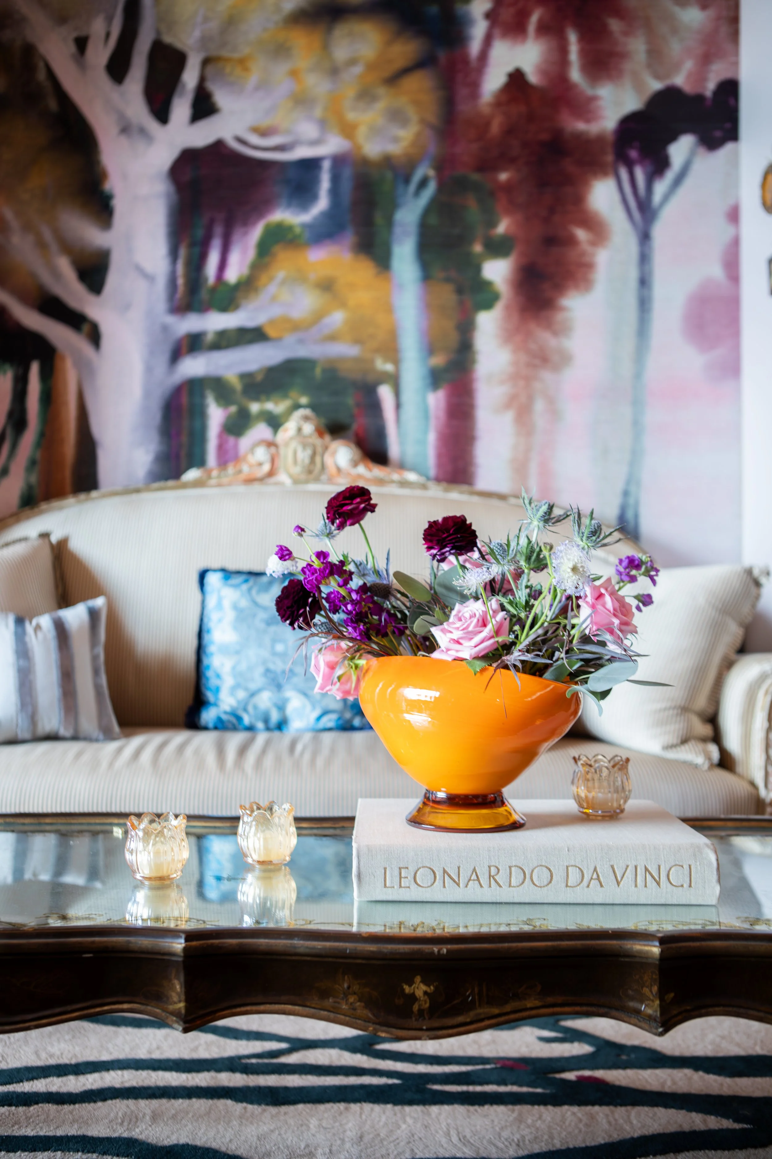 Living room with a beige striped sofa, colorful floral arrangement in an orange vase on a large book titled "Leonardo Da Vinci," and a painted abstract mural of trees on the wall behind.