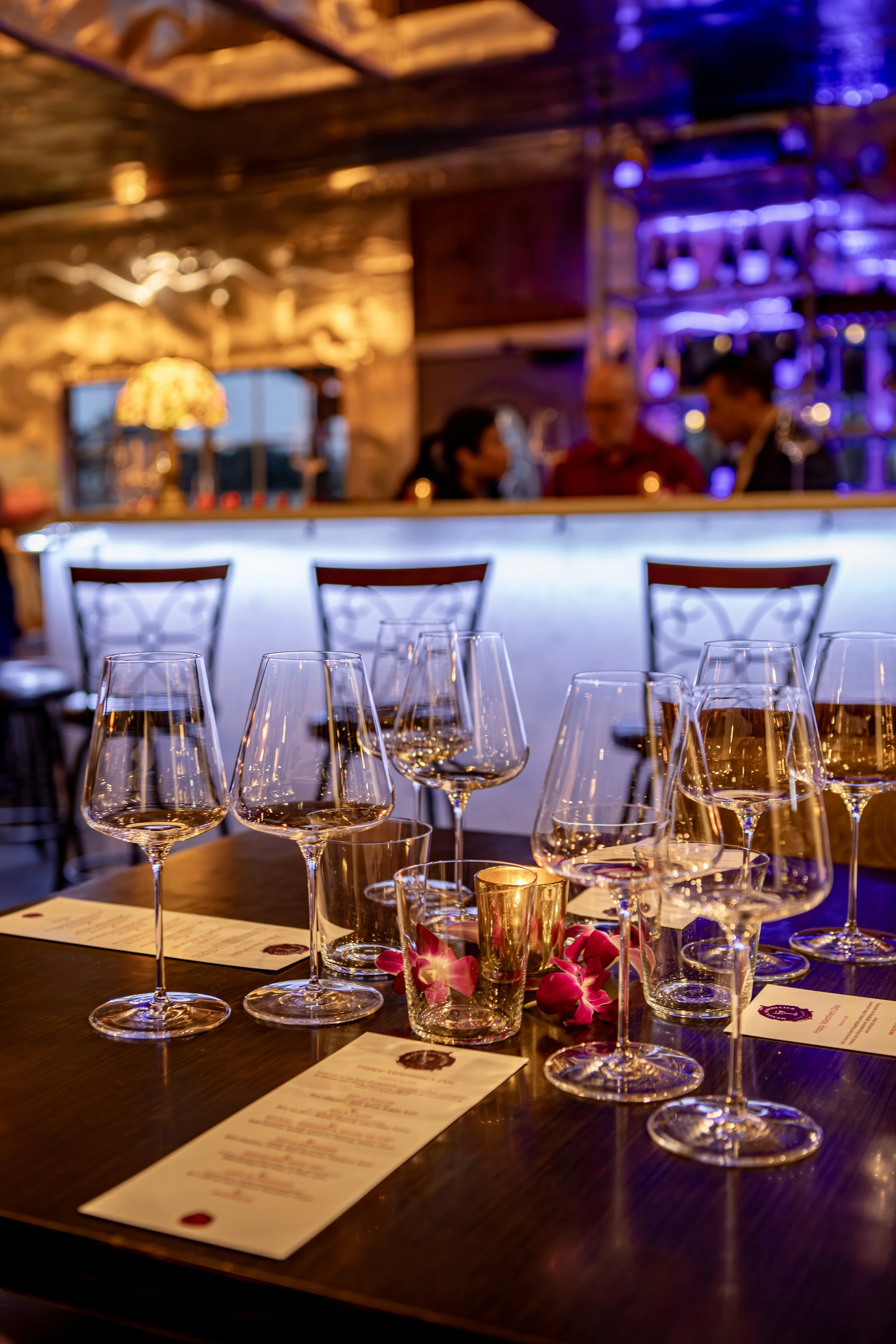 A table set with multiple wine glasses, pink flowers, and candle holders in a dimly lit restaurant or bar with a bar counter and people in the background.