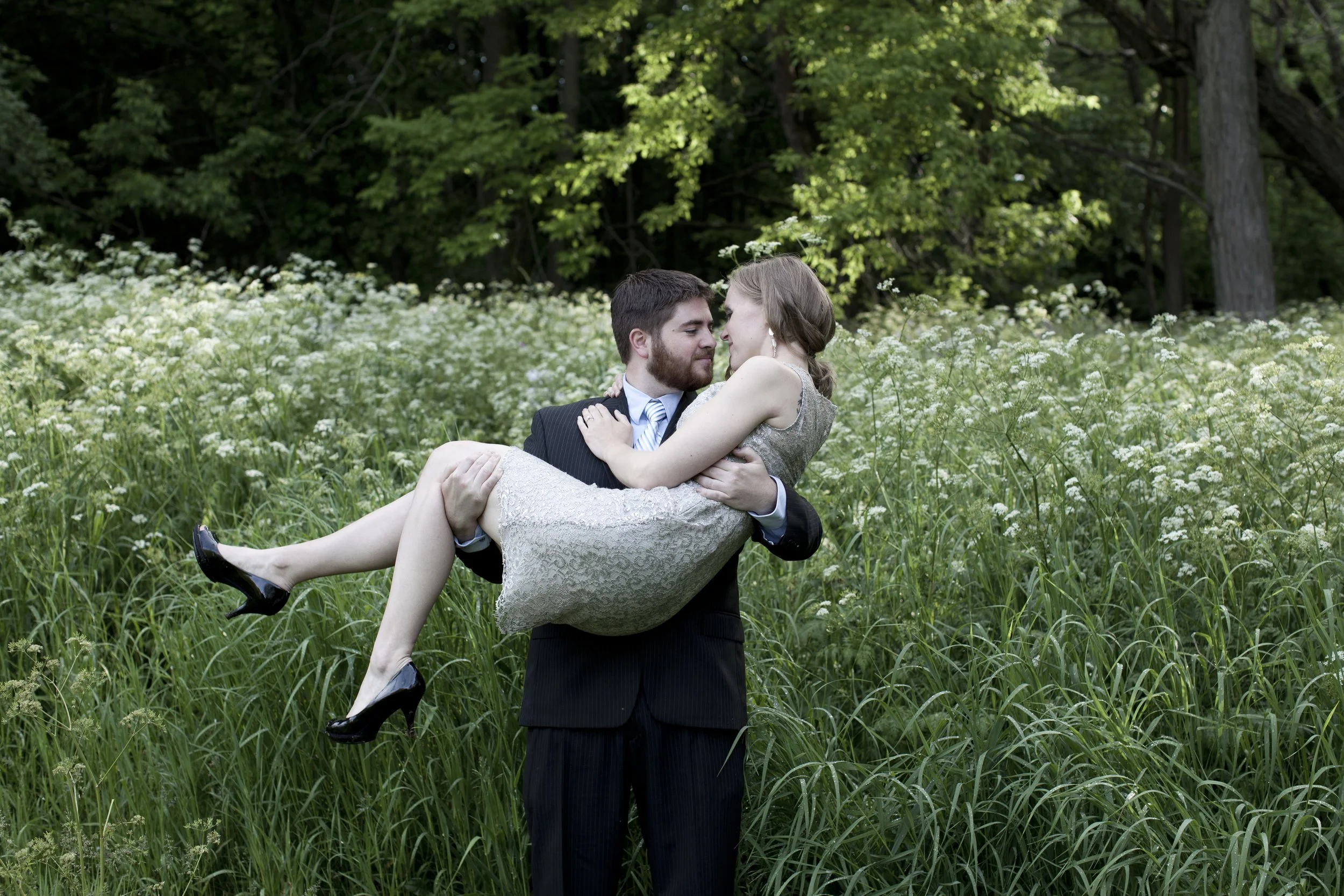 A man in a suit holding a woman in a dress in a field of tall grass and white flowers, with trees in the background. Photographed by Houston Photographer Katie Park.