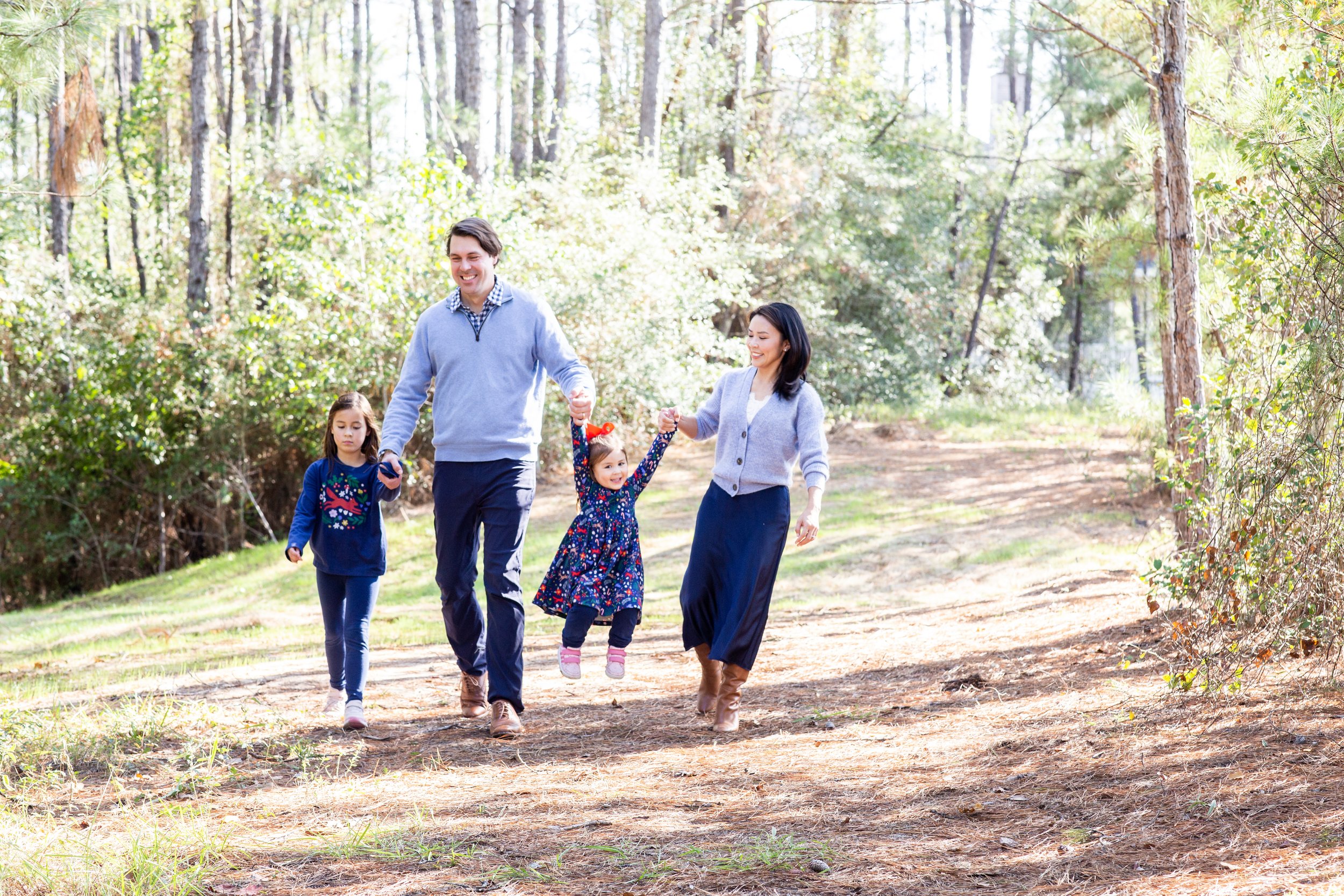 A family of four walking on a forest trail during the daytime, with two children, a boy and a girl, playing and smiling while holding hands with their parents. Photographed by Houston Photographer Katie Park.