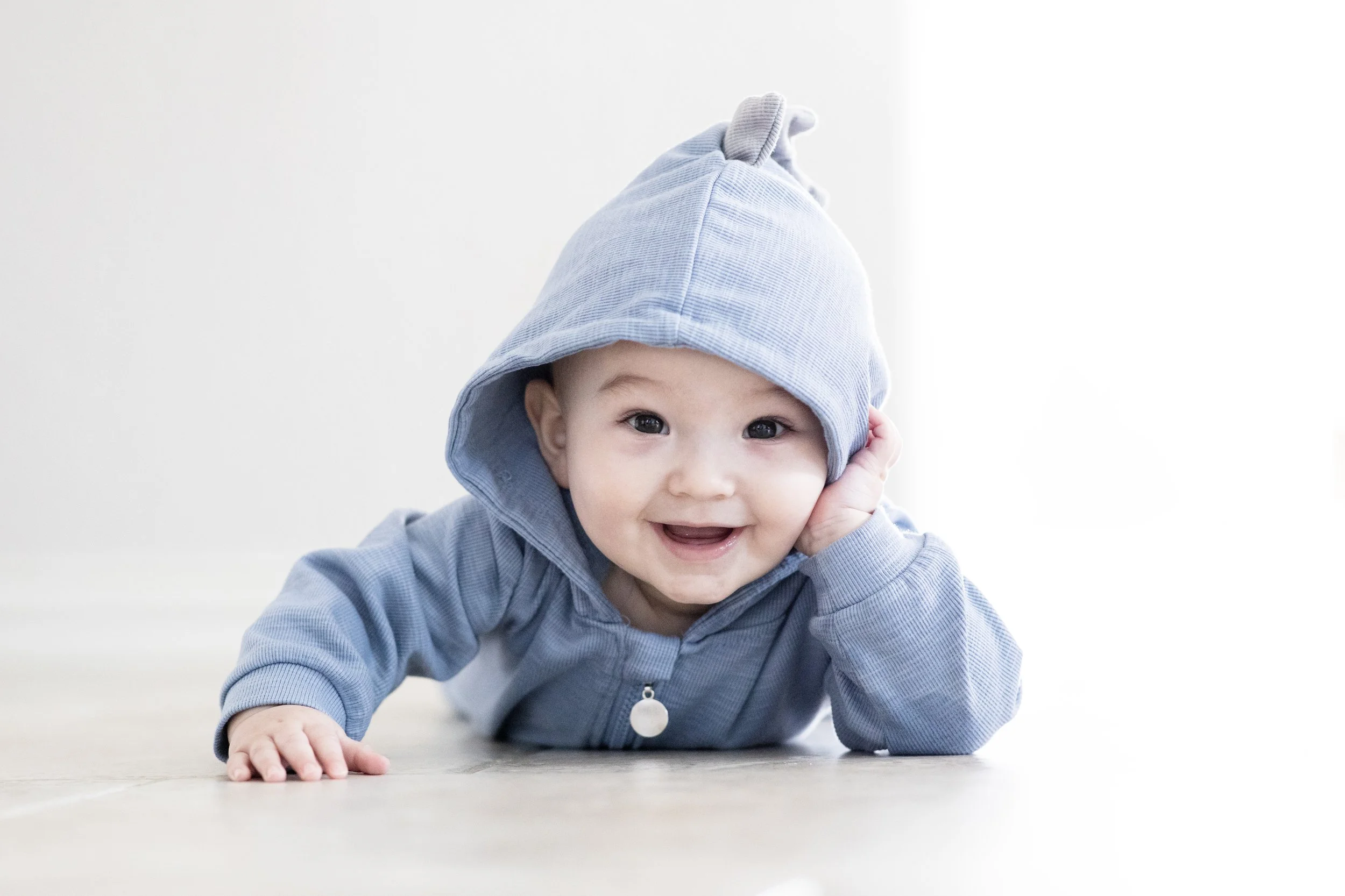 Smiling baby lying on the floor wearing a blue hoodie with a cute animal design, resting their head on their hand. Photographed by Houston Photographer Katie Park.