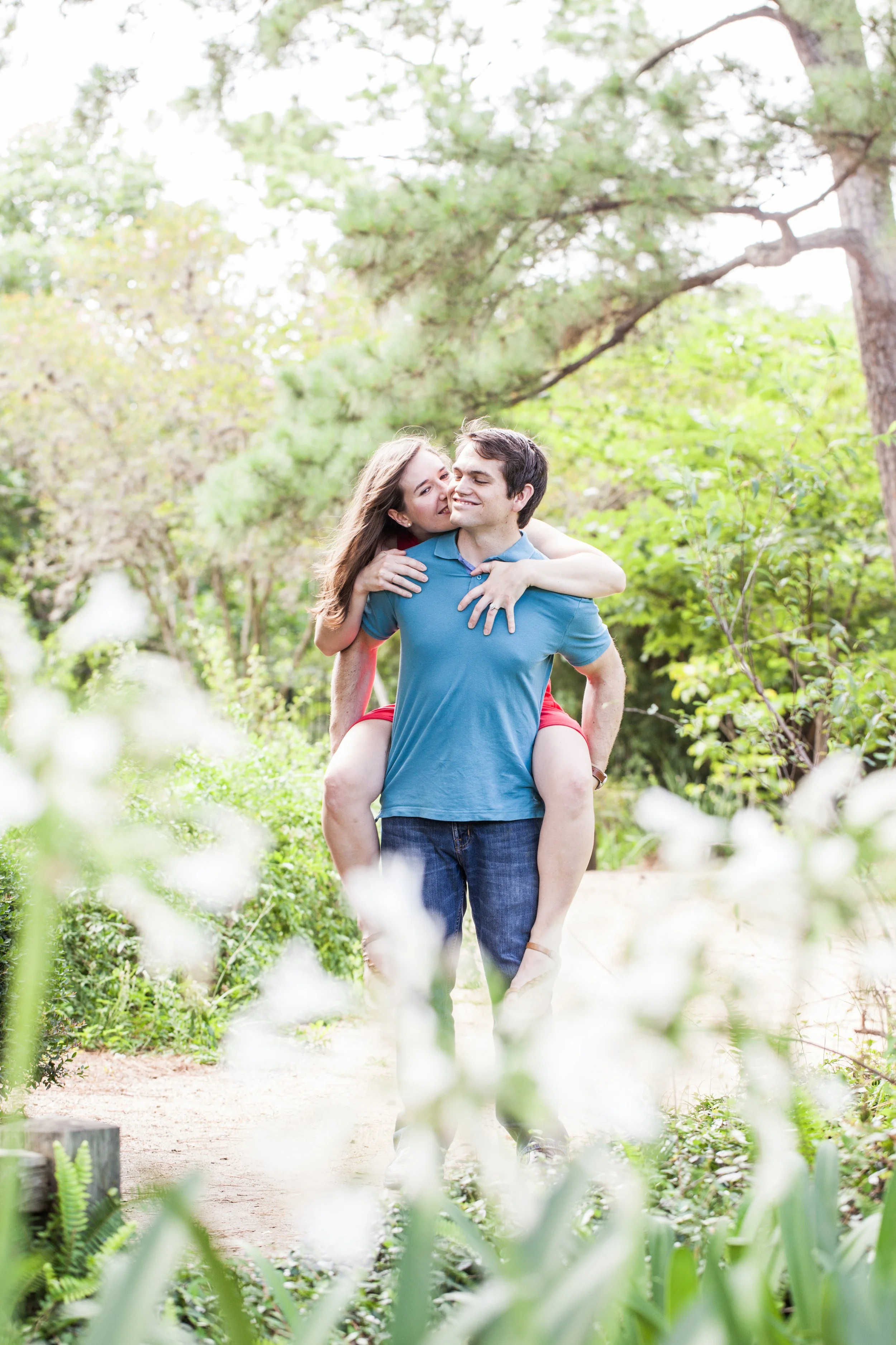 A young couple in casual clothing, with the woman in a red dress, enjoying a playful moment outdoors in a lush, green park. Engagement session Houston. Photographed by Houston Photographer Katie Park.