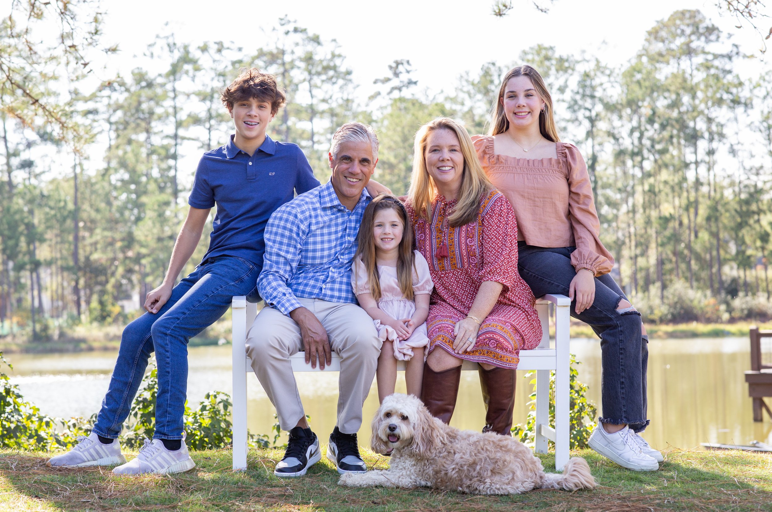 A family of six with three children, two adults, and a dog posing outdoors near a lake or river, surrounded by trees and greenery. Photographed by Houston Photographer Katie Park.