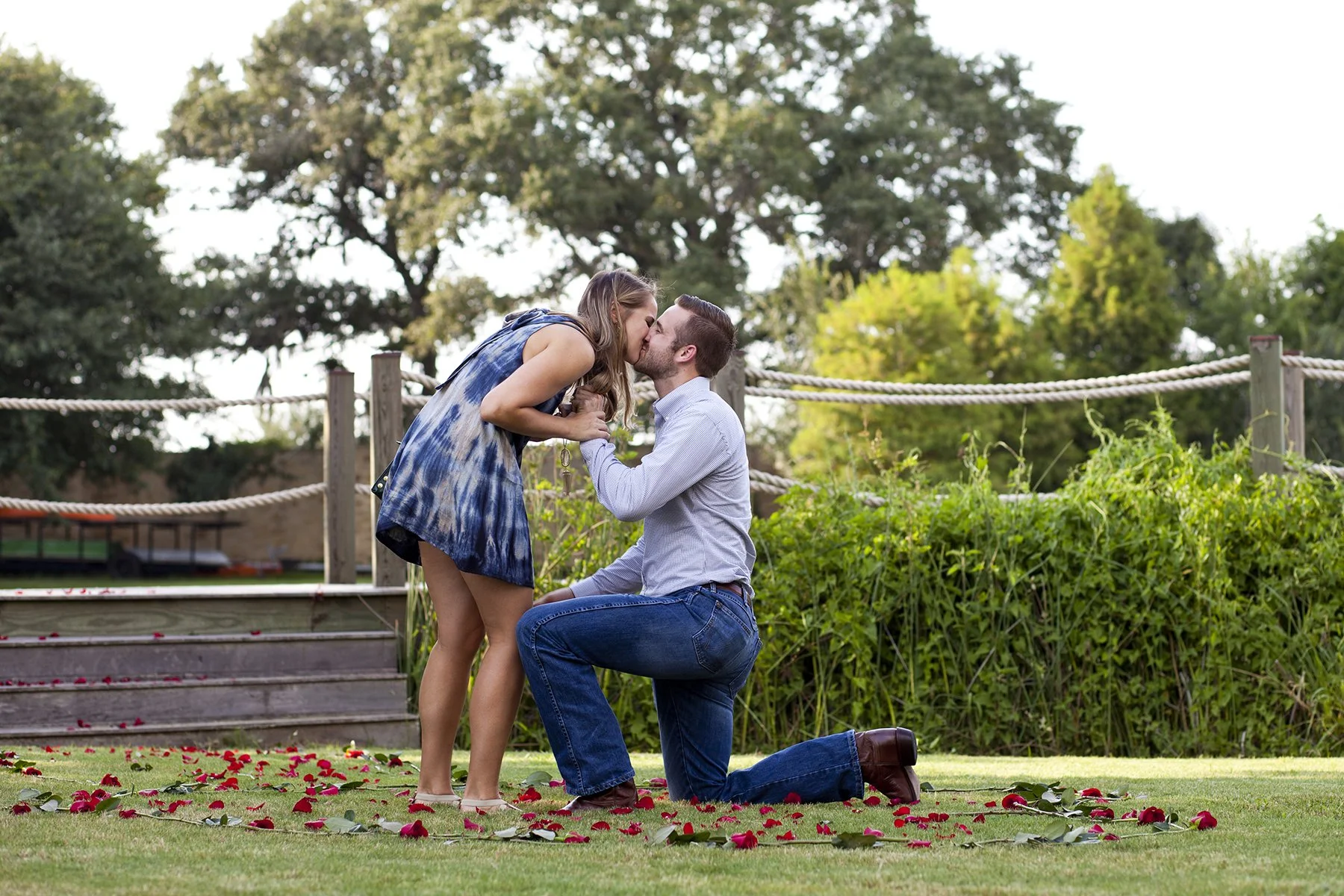 A man proposing marriage to a woman on one knee in a park, with red flower petals on the grass and greenery in the background. Photographed by Houston Photographer Katie Park.