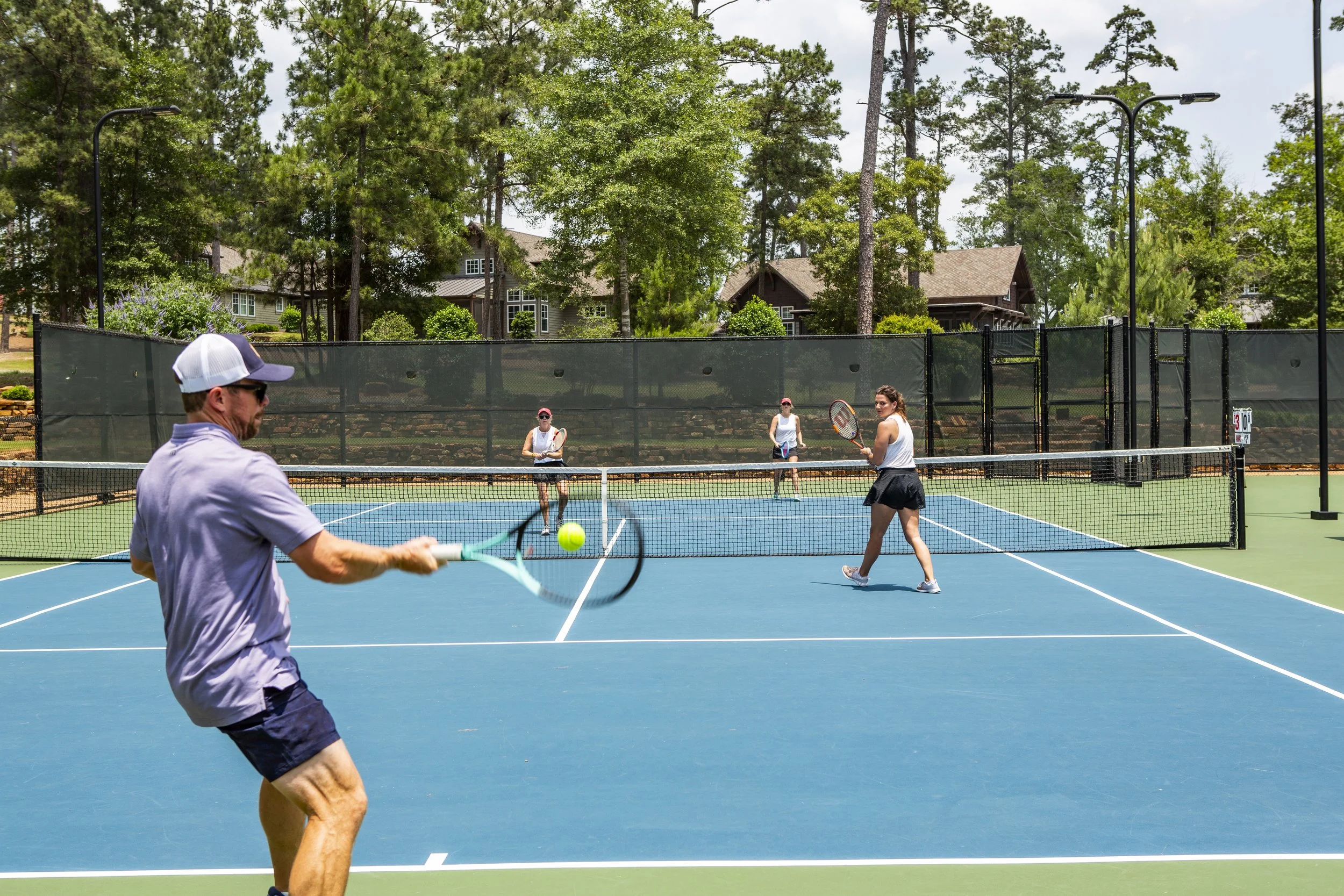 People playing pickleball on an outdoor court surrounded by trees and houses.