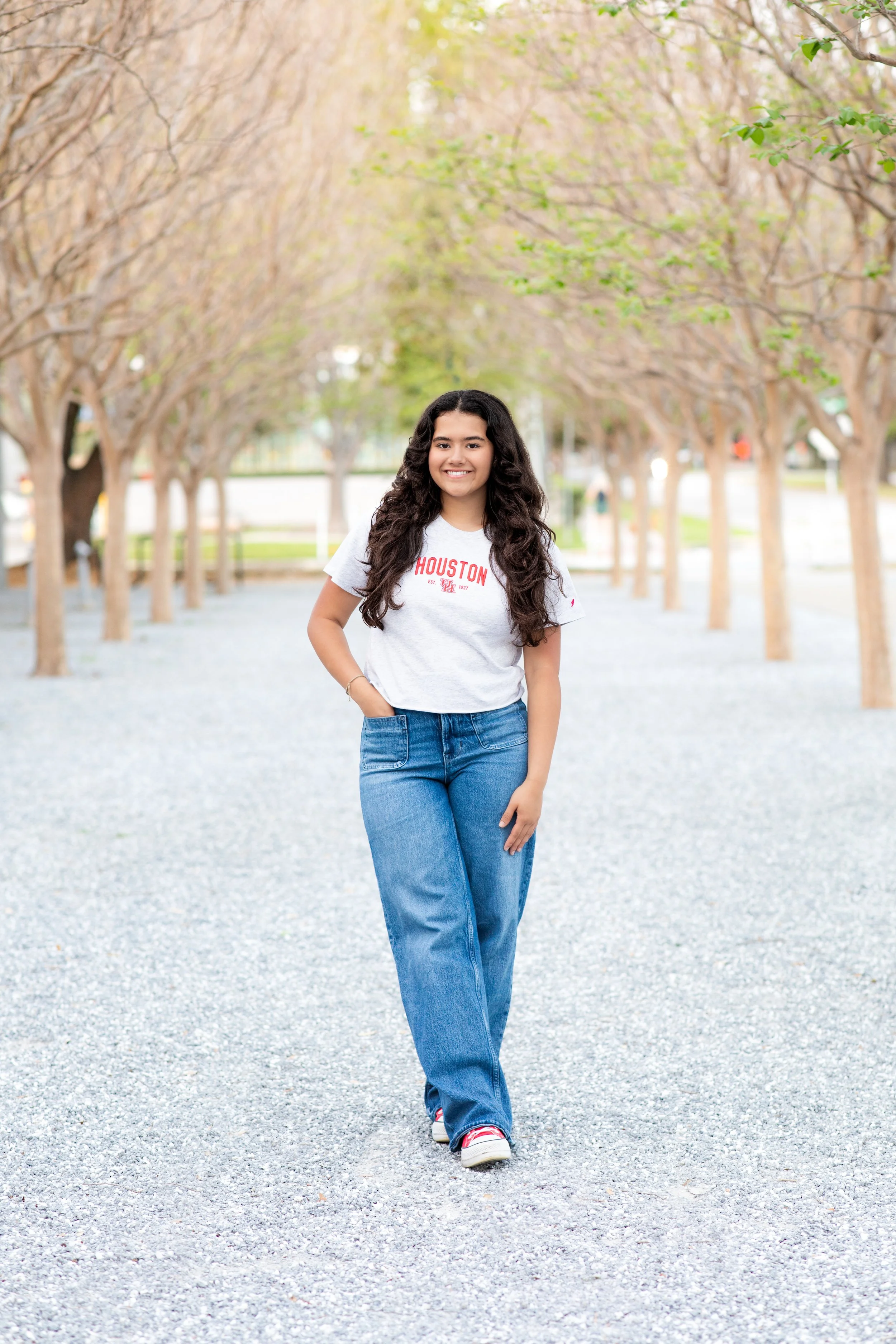 A young woman walking outdoors on a gravel path lined with evenly spaced trees with budding leaves, smiling at the camera. Photographed by Houston Photographer Katie Park.
