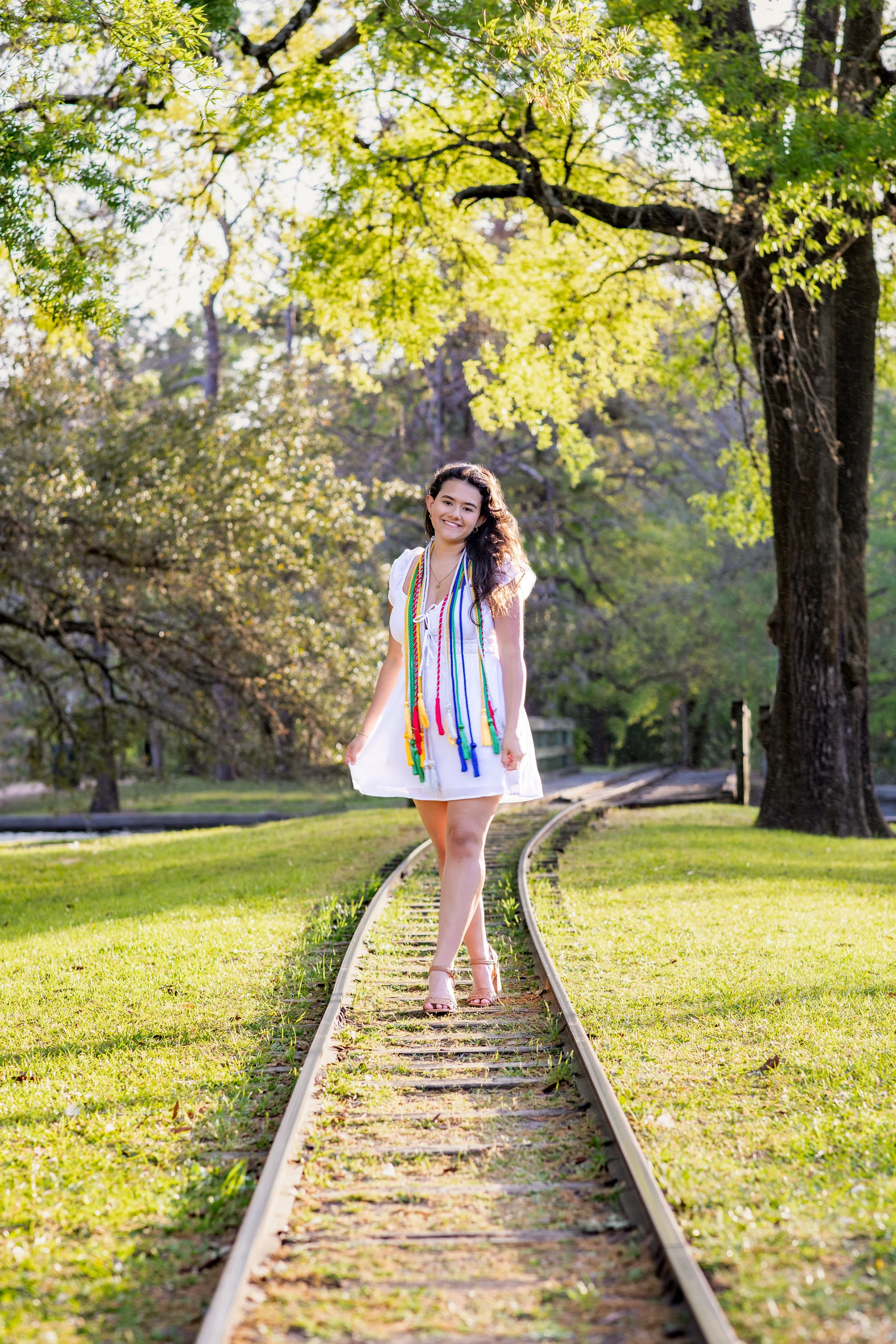 A young woman in a white dress with colorful necklaces standing on a narrow train track in a park with green trees and sunlight. Photographed by Houston Photographer Katie Park.