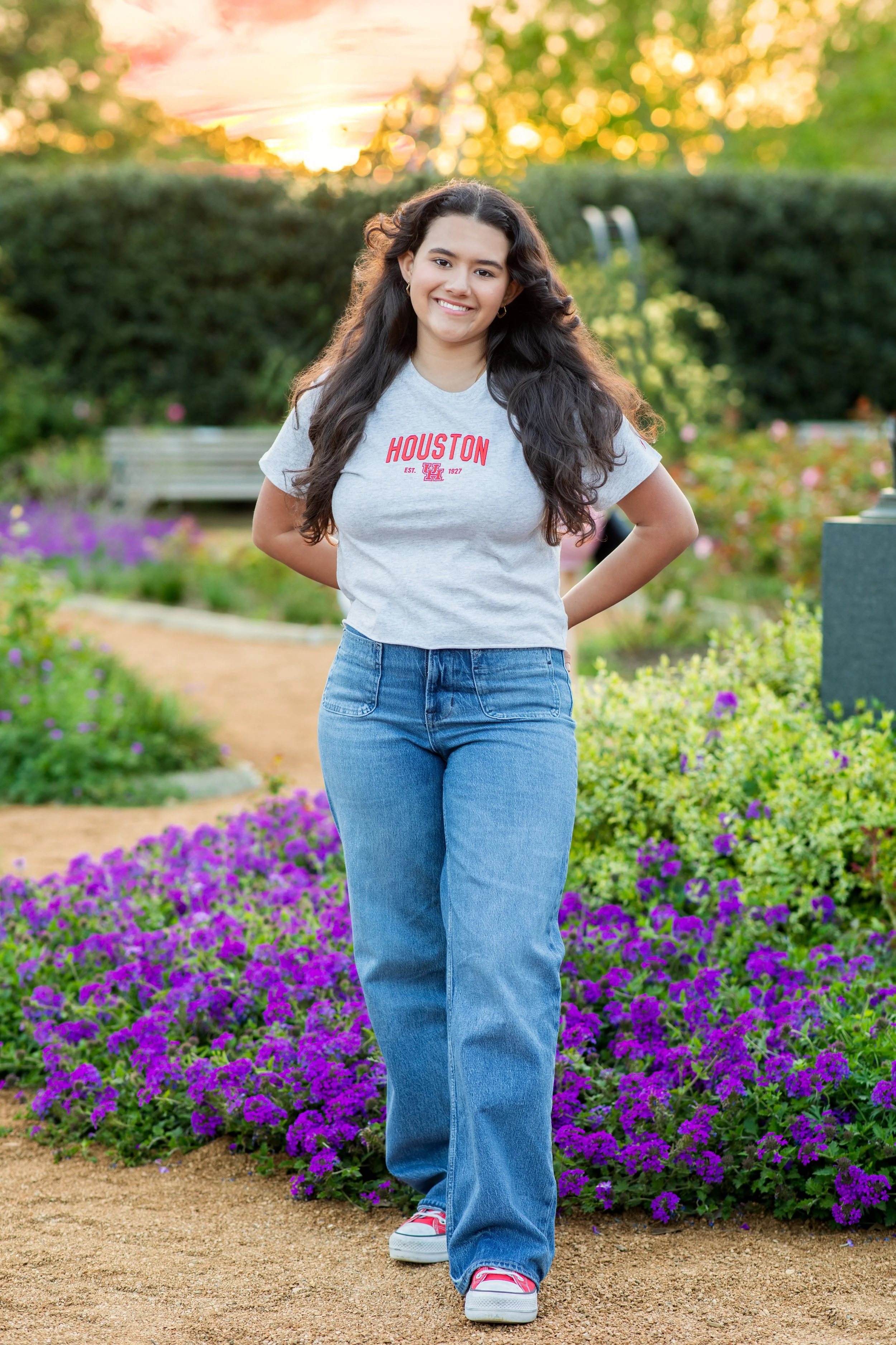 Young woman with long dark hair, wearing a gray Texas Houston t-shirt, blue jeans, and sneakers, standing in a garden with purple flowers and a sunset in the background. Photographed by Houston Photographer Katie Park.