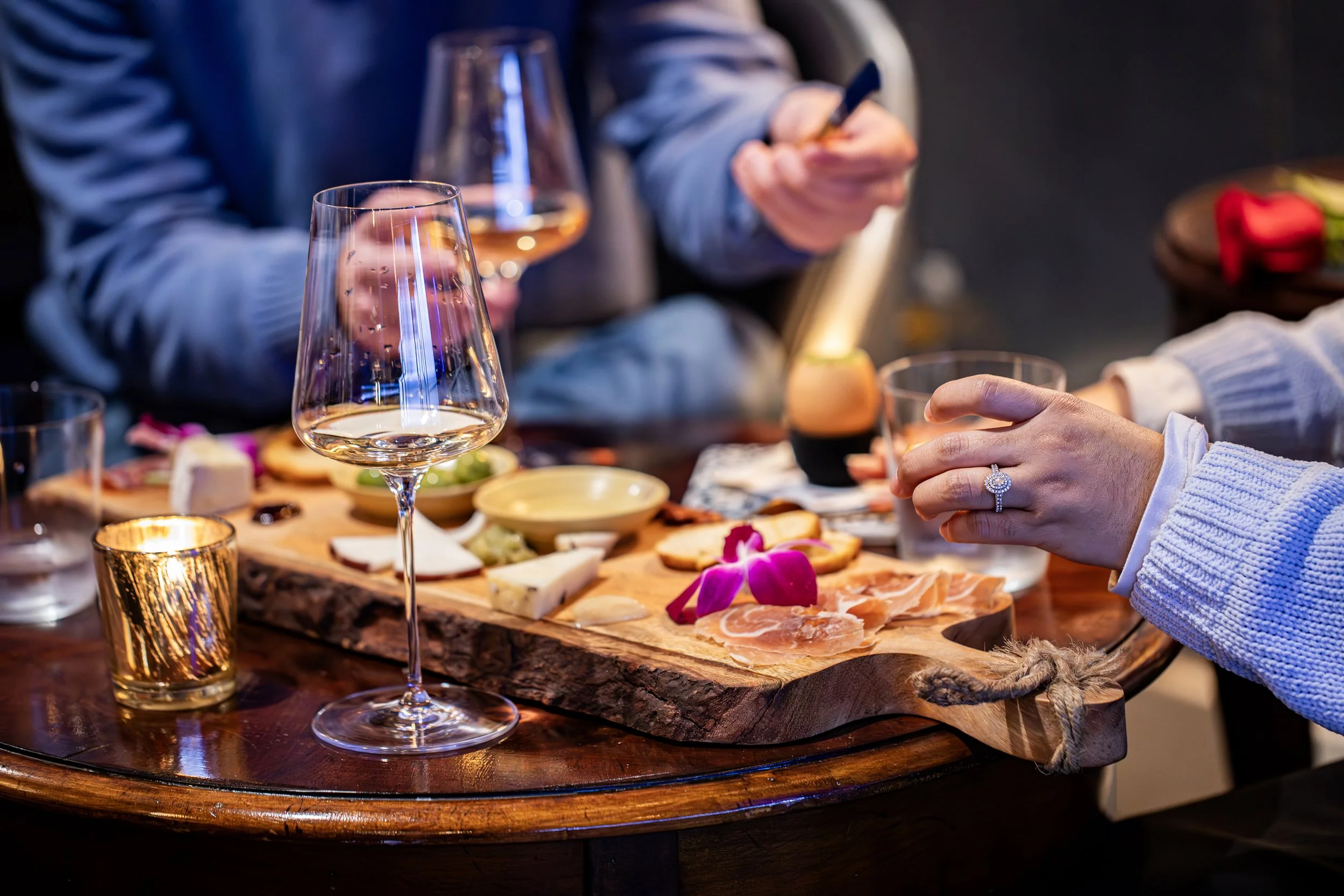 A charcuterie board with various cheeses, cured meats, and flowers on a wooden table, with wine glasses and a candle, in a cozy setting.