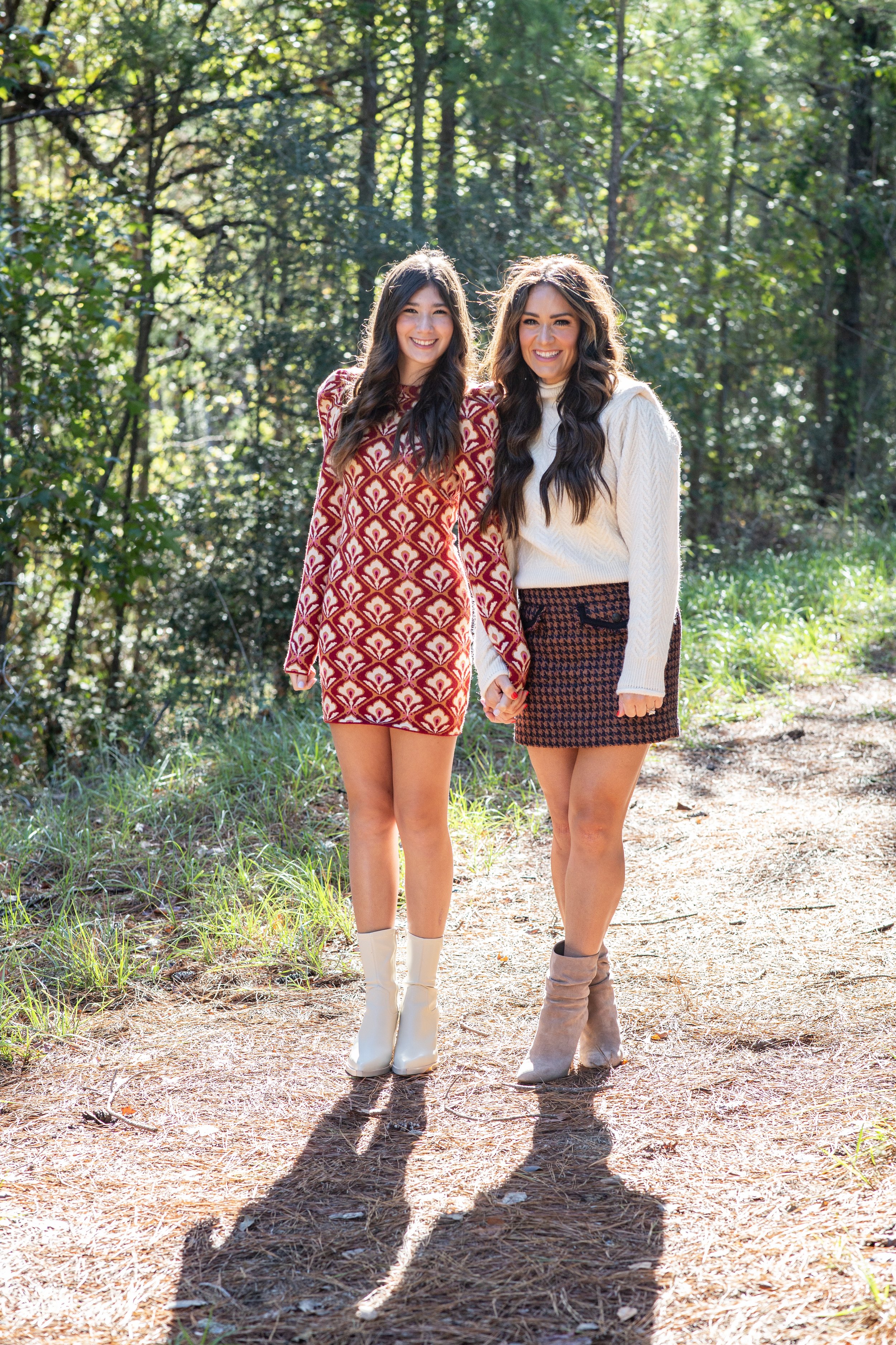 Two young women walking hand-in-hand on a forest trail during daytime, smiling happily, dressed in stylish clothes, sunlight filtering through trees. Photographed by Houston Photographer Katie Park.