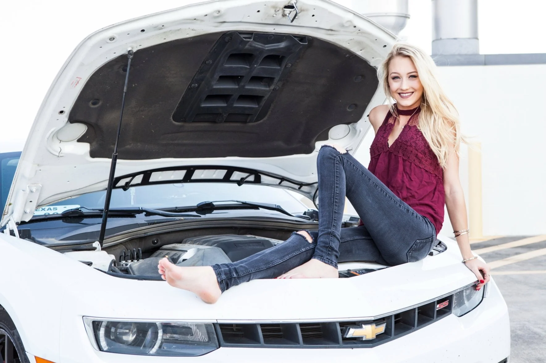 A young woman with blonde hair sitting on the hood of a white Chevrolet car with the hood open, smiling at the camera. She is wearing a maroon sleeveless top and dark jeans. Photographed by Houston Photographer Katie Park.