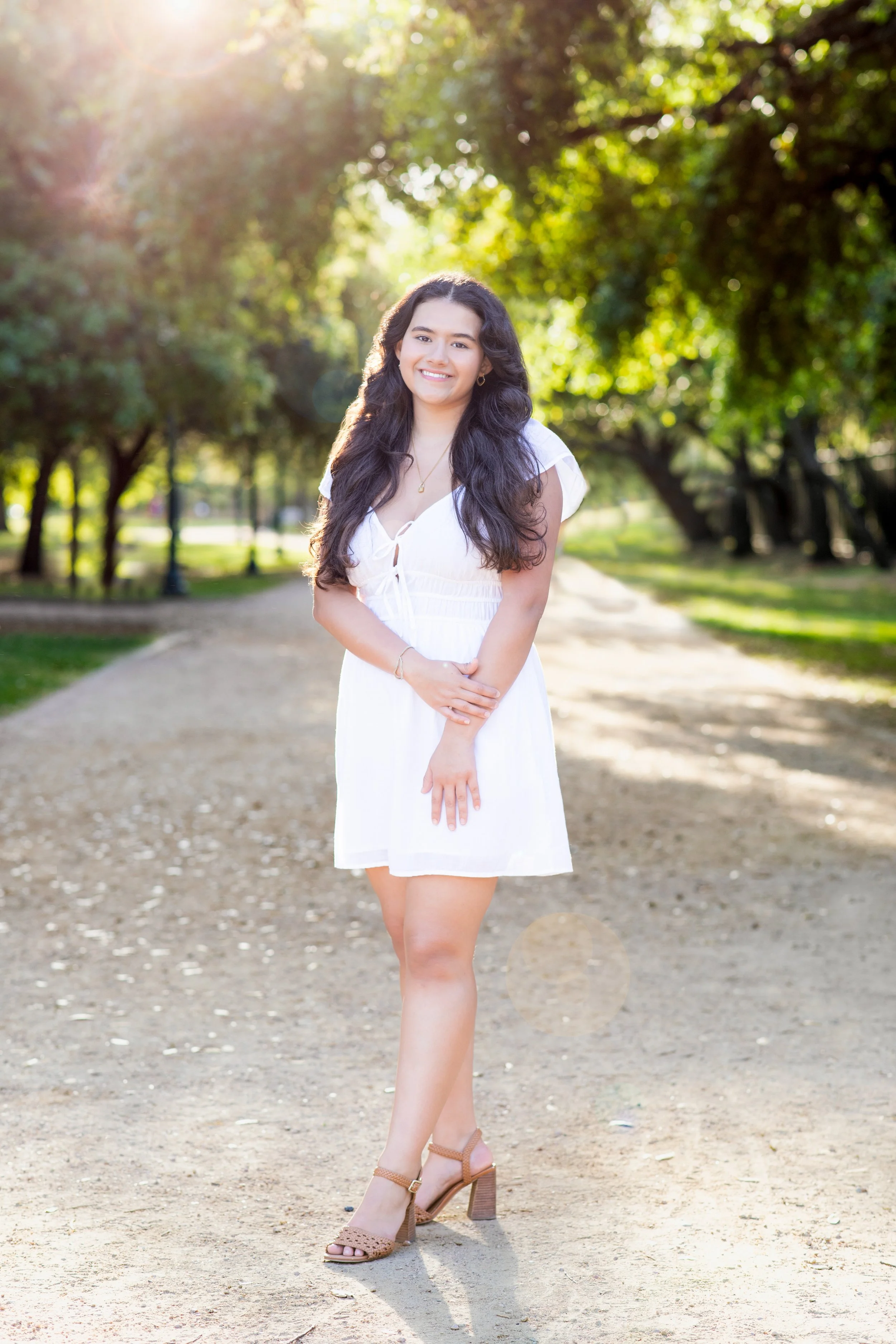 A young woman with long dark hair wearing a white dress and tan high-heeled sandals standing on a dirt path in a park with lush green trees in the background, sunlight shining through the trees. Photographed by Houston Photographer Katie Park.