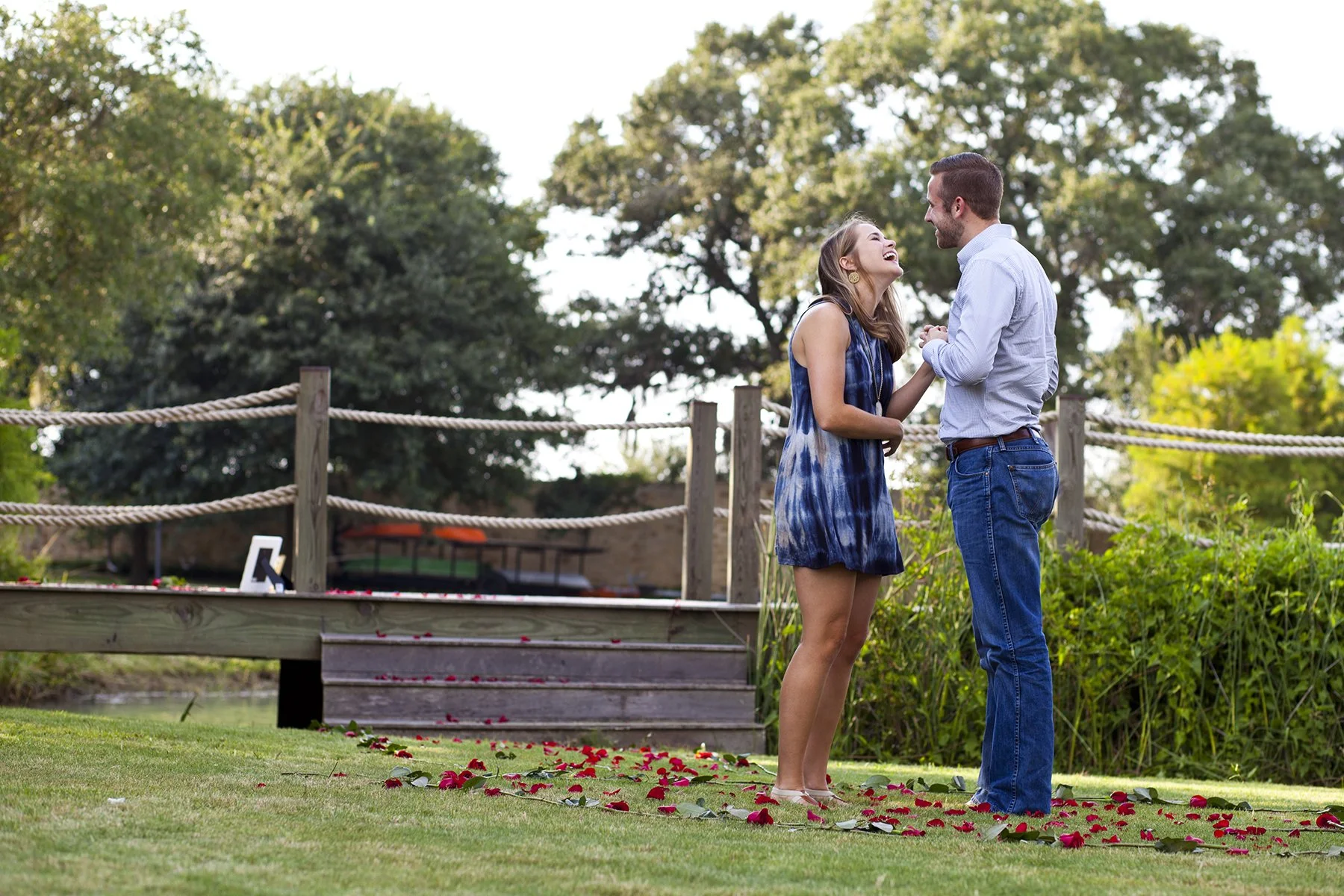 A man and woman stand outdoors on a grassy area with red flower petals scattered on the ground. They are holding hands, smiling, and looking at each other in a joyful moment. Trees and a wooden railing are in the background. Photographed by Houston P