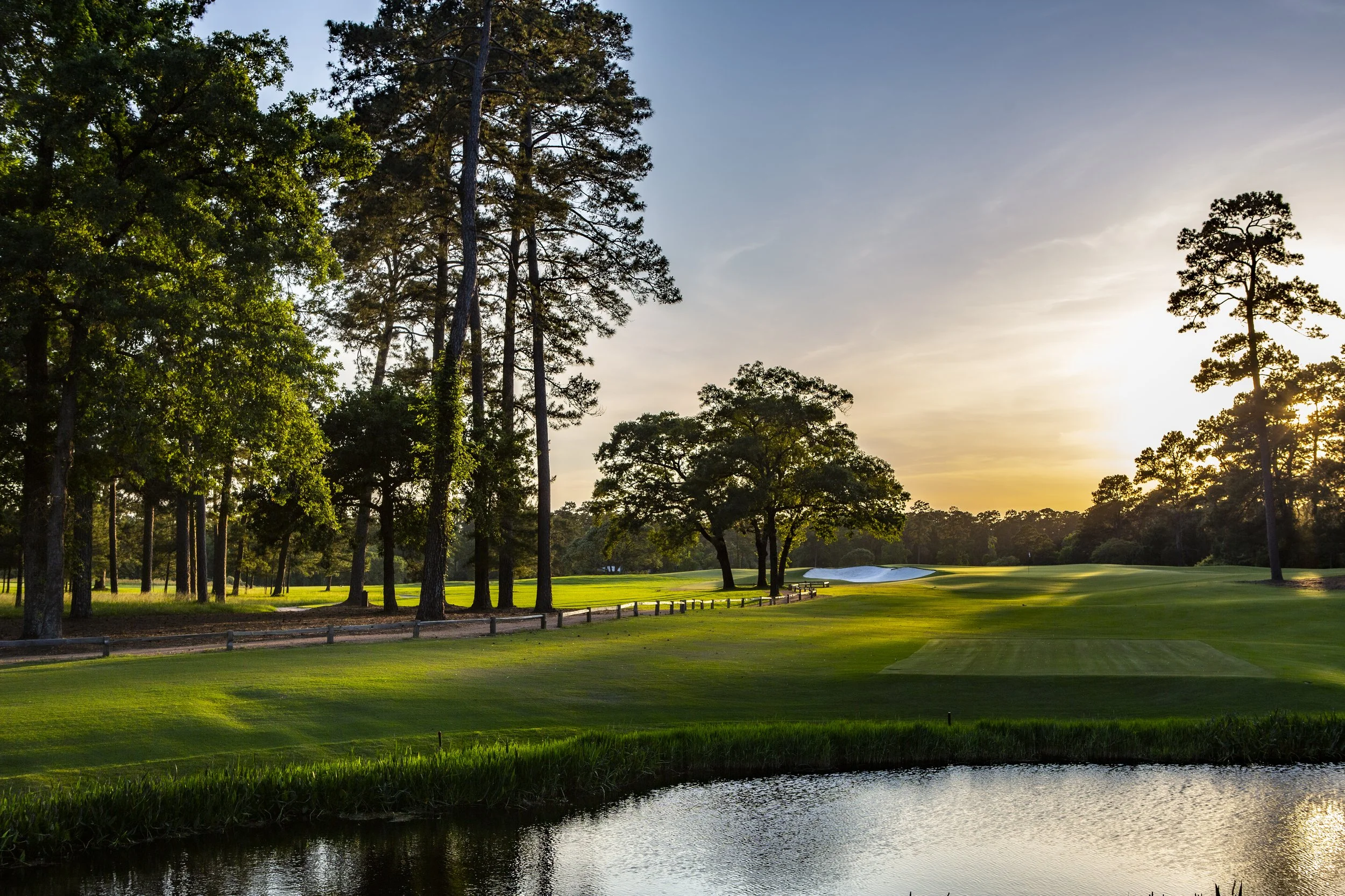 A scenic view of a golf course during sunset, featuring lush green grass, trees lining the course, a water hazard in the foreground, and a sand trap near the green.
