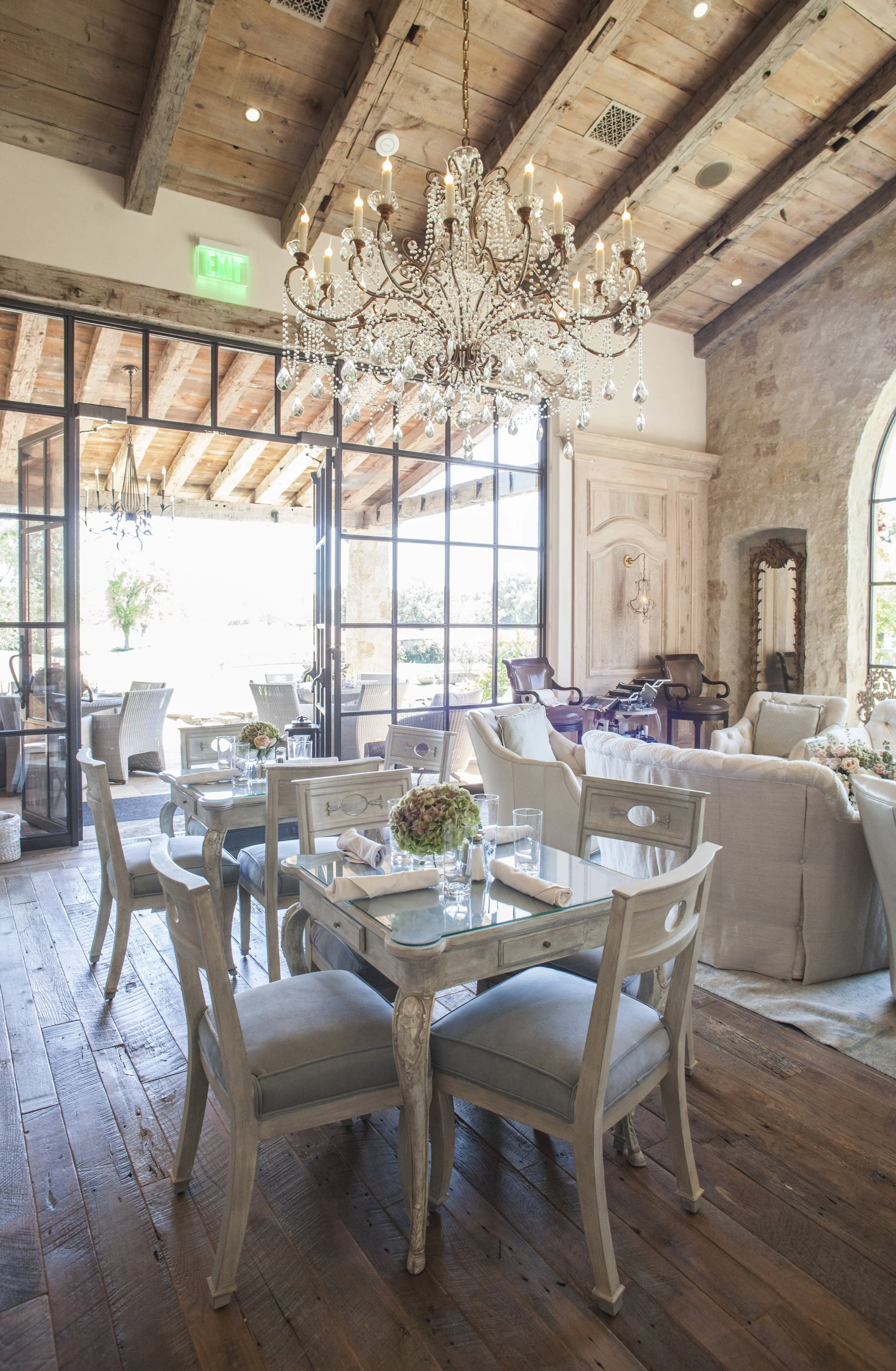 Elegant dining room with rustic wooden floors, a large crystal chandelier, and a mix of vintage and modern furniture, including a glass-top table with floral arrangements and cushioned chairs.